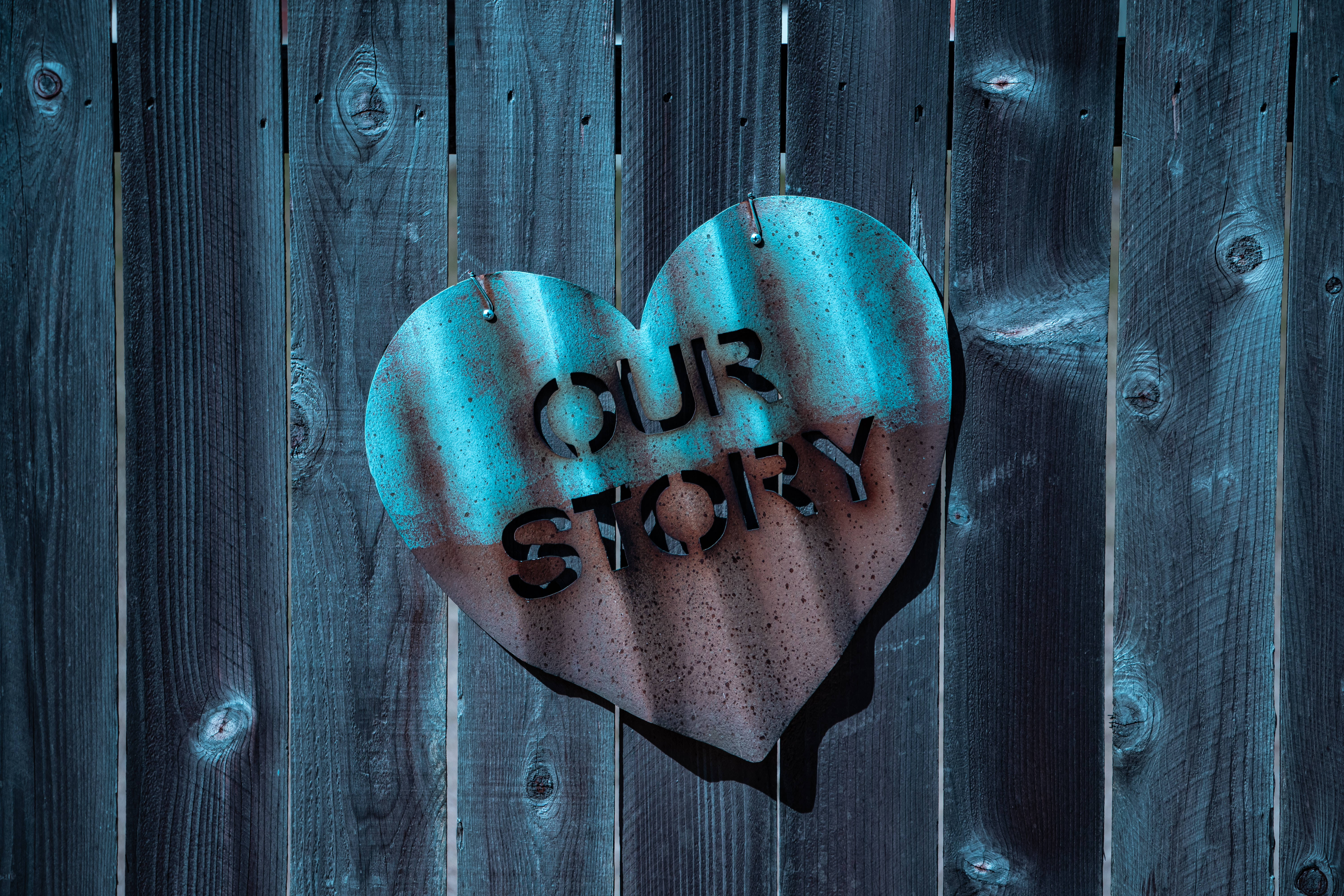 Love Story 桌布 壁紙 — 一個風化的心形金屬標牌，上面寫著「OUR STORY」，掛在帶有鏽跡和藍色銅綠的深色木柵欄上。