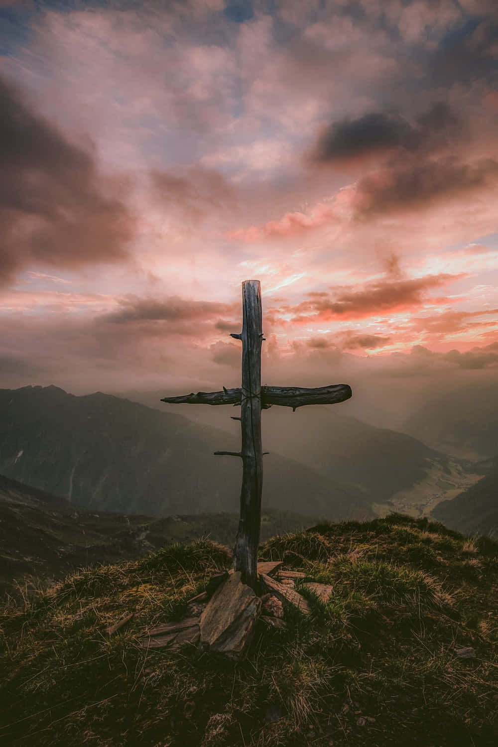 Cool Cross 桌布 壁紙 — 一個風化的木製十字架矗立在長滿草的山脊上，背景是壯麗的日落天空，帶有粉紅色和紫色的雲彩。