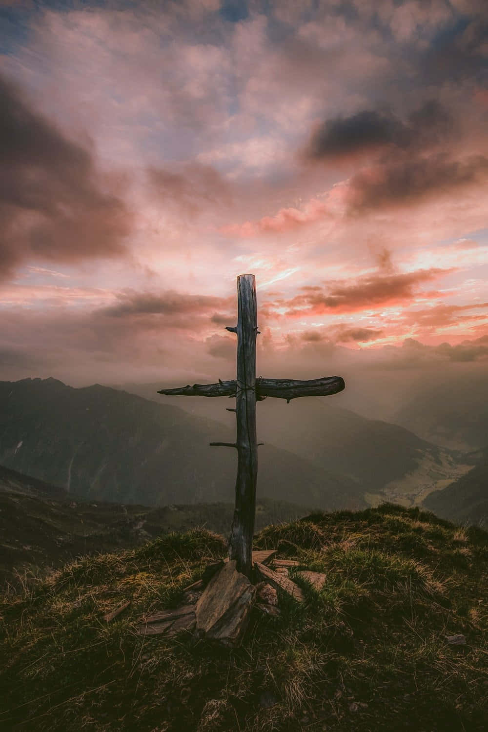 Cute Cross 桌布 壁紙 — 一個風化的木製十字架矗立在長滿草的山脊上，天空是戲劇性的日落，有粉紅色和紫色的雲彩。