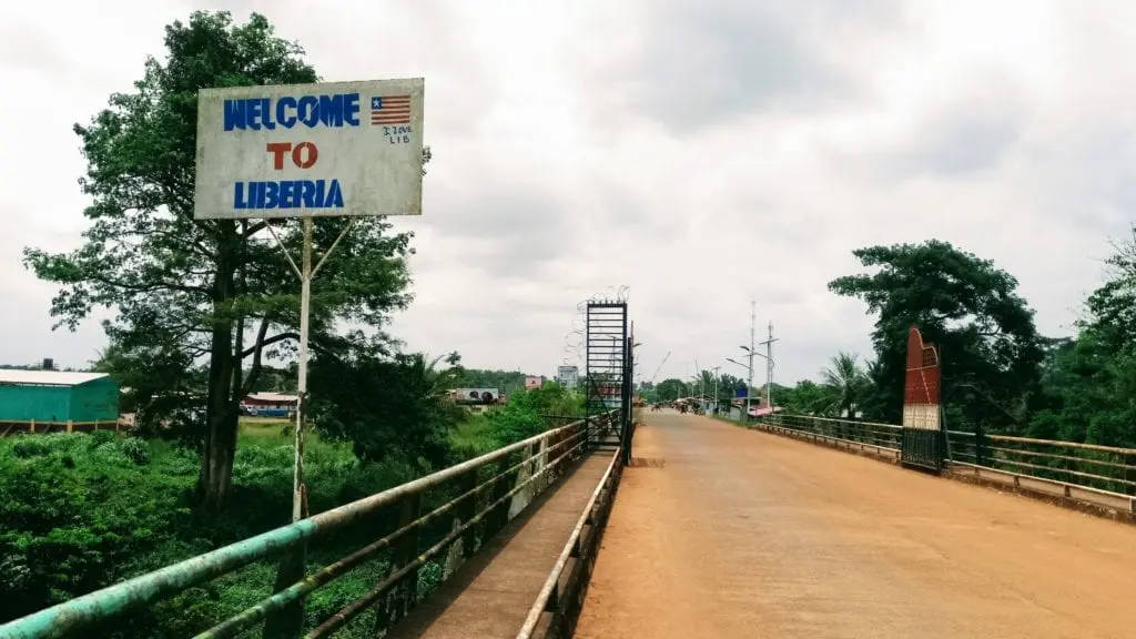 Liberia 桌布 壁紙 — 一個風化的歡迎來到利比里亞標誌，站在一座生鏽的金屬橋旁邊，橋橫跨鬱鬱蔥蔥的綠色景觀，天空多雲。