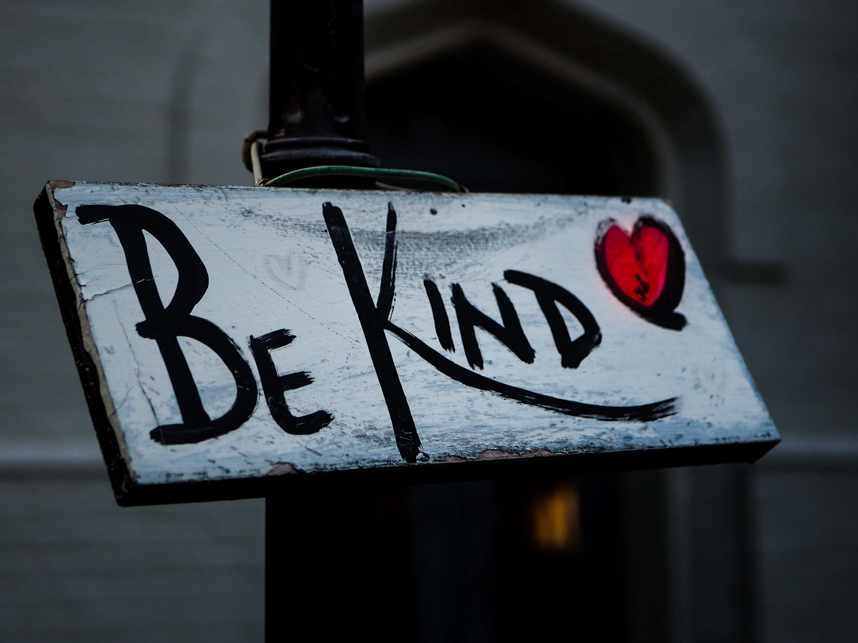 Be Kind 桌布 壁紙 — 一個風化的白色標牌，上面有粗體黑色塗鴉寫著「Be Kind」，旁邊有一個紅色愛心，安裝在柱子上，背景是昏暗的城市景觀。