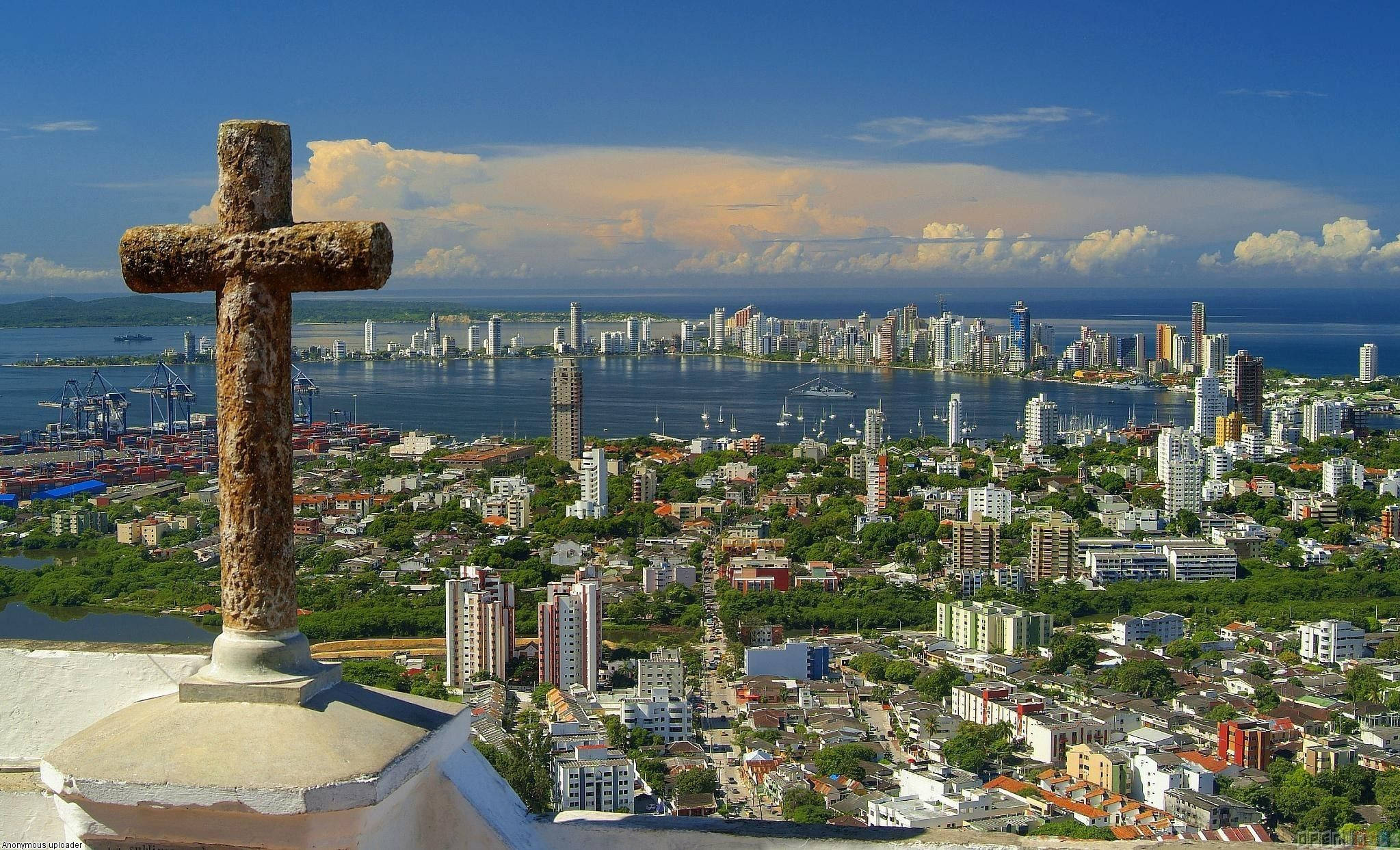 Colombia 桌布 壁紙 — 一個風化的石十字架俯瞰著一個廣闊的海濱城市，擁有現代摩天大樓和港口，天空湛藍，雲彩壯麗。