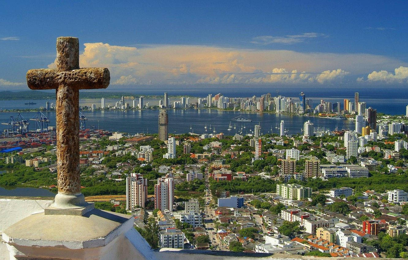 Colombia 桌布 壁紙 — 一個風化的石十字架俯瞰著一座沿海城市，城市中有現代高樓大廈和繁忙的港口，天空壯麗。