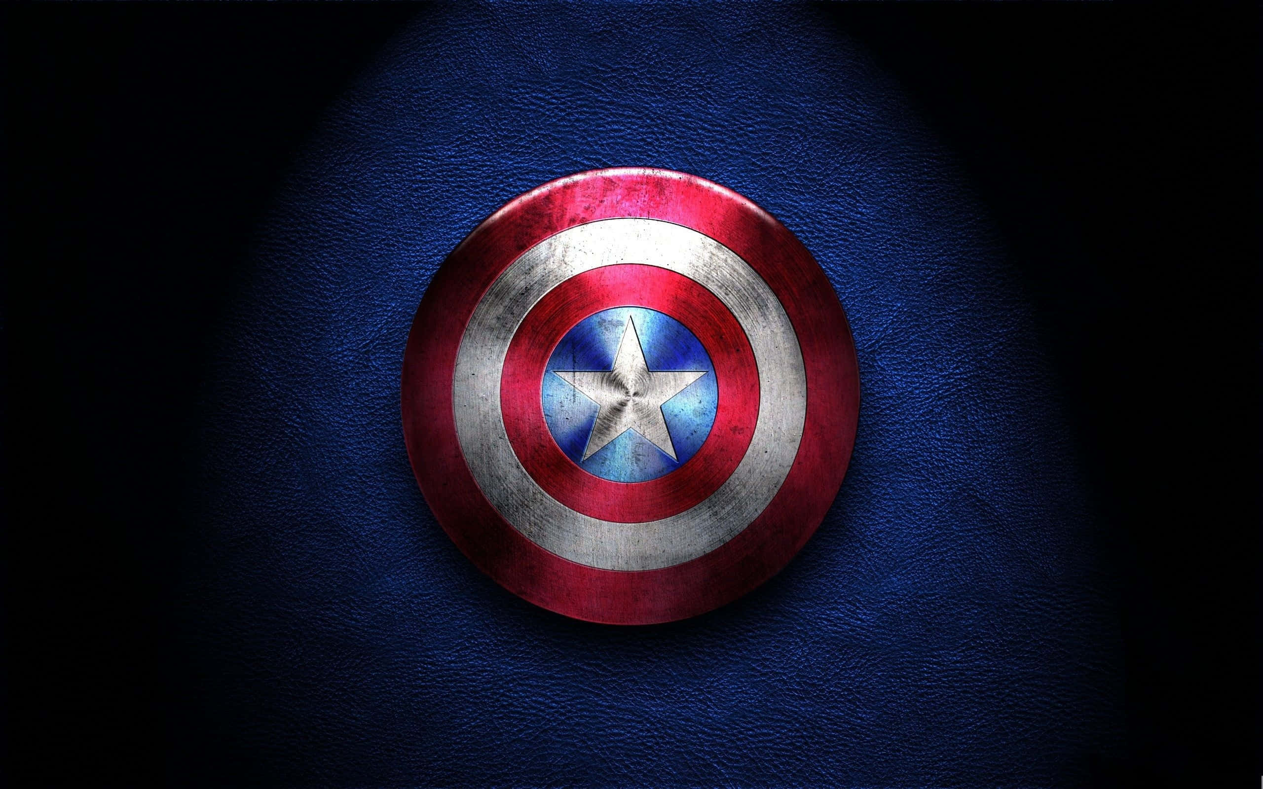 Captain America Logo 桌布 壁紙 — 一個風化的美國隊長盾牌在戲劇性聚光燈下放在有紋理的藍色皮革上。