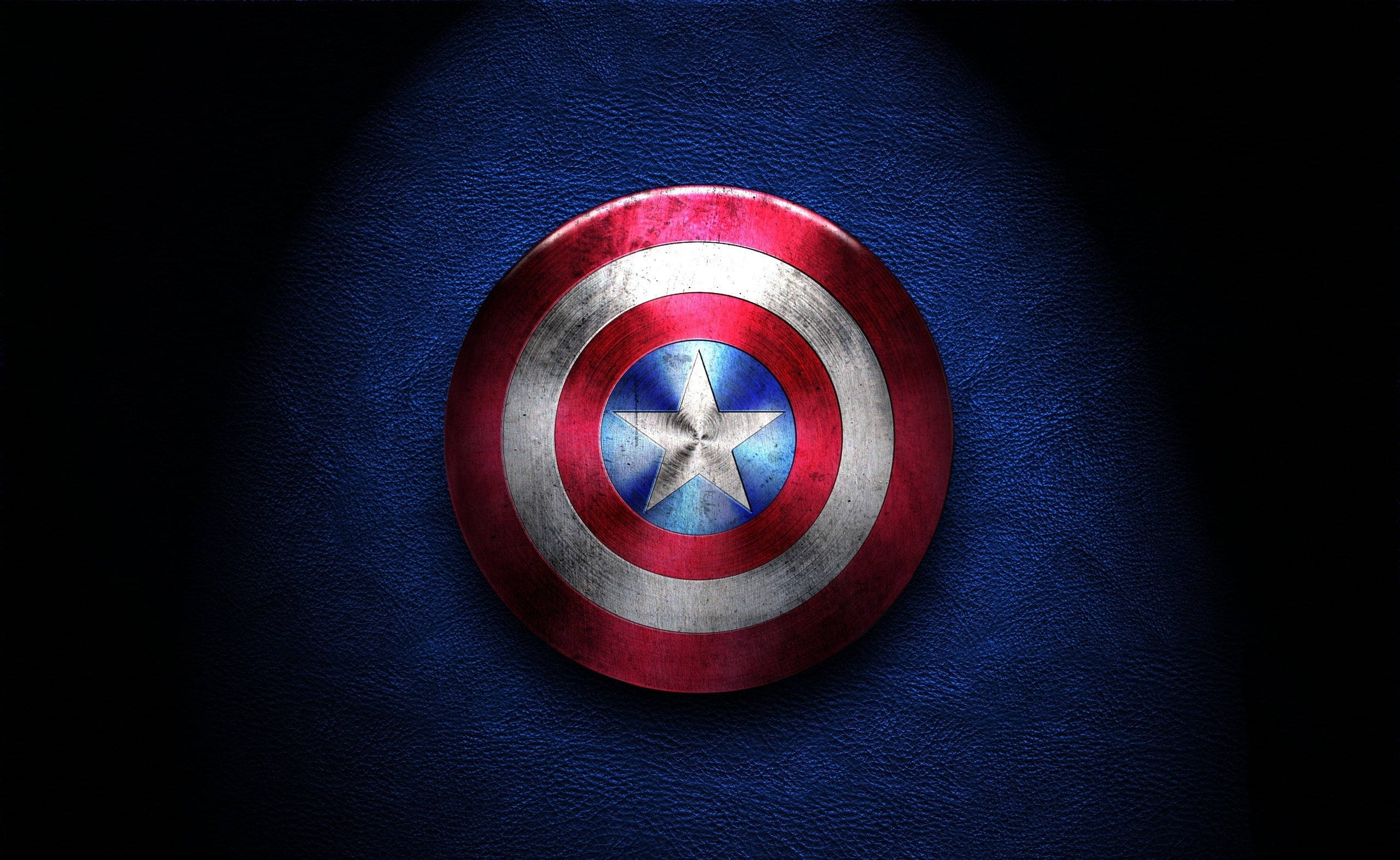Marvel Captain America 桌布 壁紙 — 一個風化的美國隊長盾牌放置在帶有紋理的藍色皮革上，在黑色背景下受到戲劇性的聚光燈照射。