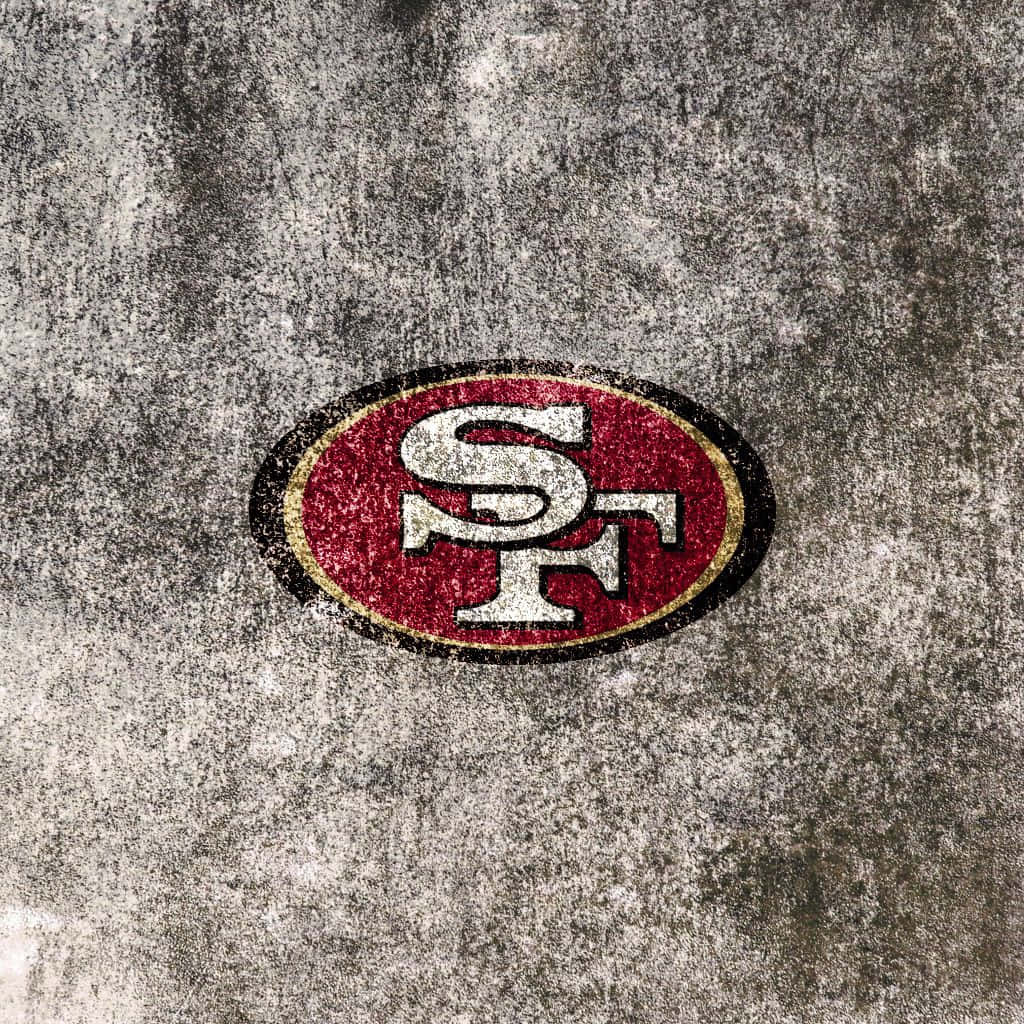 49ers 桌布 壁紙 — 一個風化的舊金山49人隊標誌，紅、白、金色在粗糙的混凝土表面上顯得褪色。
