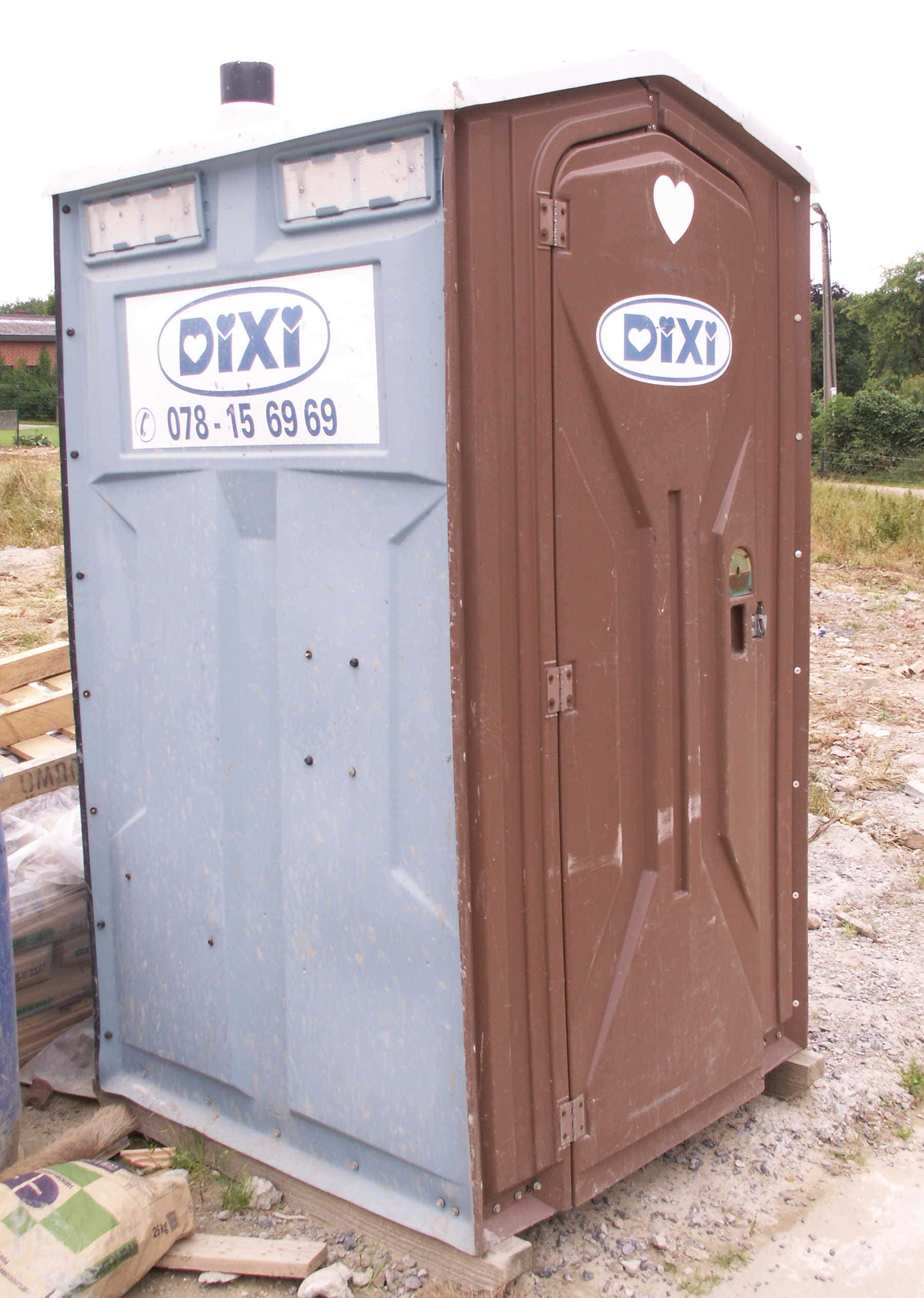Outhouse 桌布 壁紙 — 一個風化的藍色和棕色Dixi便攜式廁所，在陰天之下，站在建築材料旁邊的碎石地上。