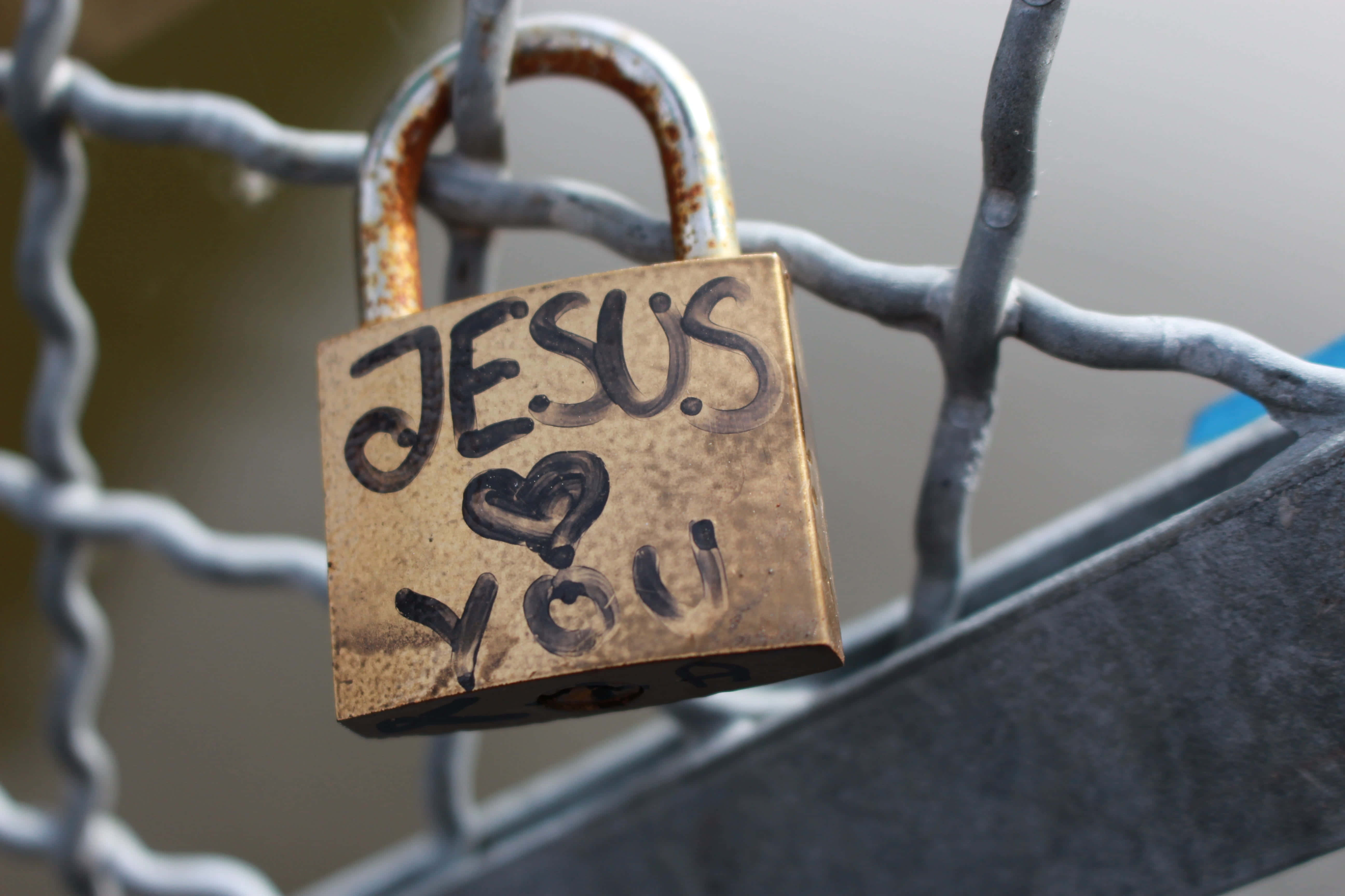 桌布 壁紙 — 一個風化的青銅掛鎖，上面刻有「JESUS ❤️ YOU」，掛在生鏽的鐵絲網圍欄上。