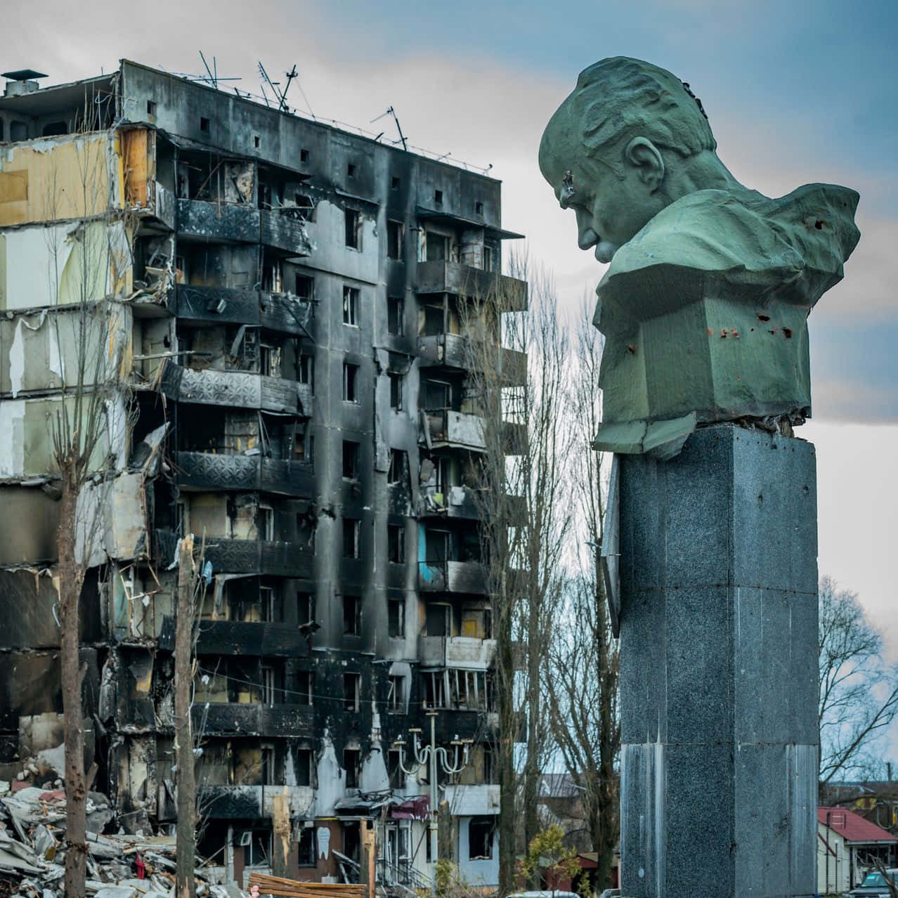 Ukraine 桌布 壁紙 — 一個風化的青銅男子半身像矗立在基座上，旁邊是一棟在多雲天空下嚴重受損且燒焦的公寓大樓。