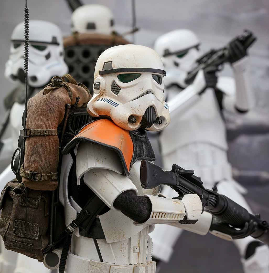 Jedha 桌布 壁紙 — 一個風化的風暴兵公仔的特寫，穿著橙色背心和背包，手持爆能槍，周圍有其他士兵在一個粗糙的場景中。