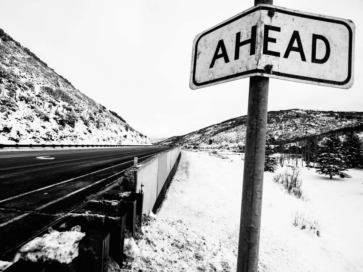 Aesthetic Black And White 桌布 壁紙 — 一個風化的黑白路牌，上面寫著AHEAD，矗立在一條灑滿雪花的公路旁，公路蜿蜒穿過一片荒涼的山區冬季景觀。