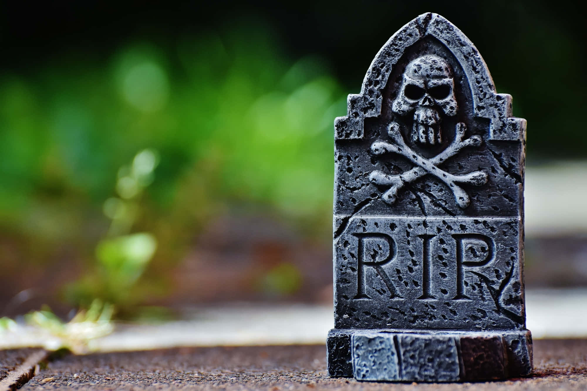 Tombstone 桌布 壁紙 — 一個風化的黑色墓碑，上面刻有骷髏頭和交叉骨圖案，放置在木質表面上，背景是模糊的綠色。
