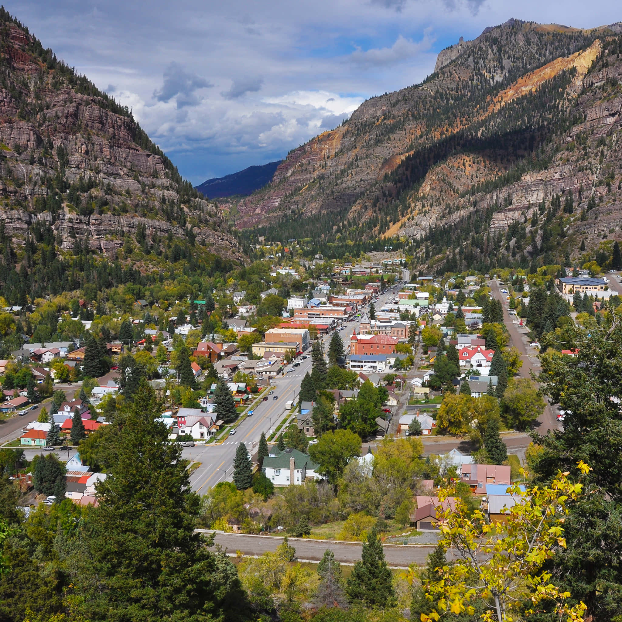 Colorado 桌布 壁紙 — 一個風景如畫的山城，坐落在山谷中，四周是陡峭的岩石懸崖和秋色樹木，天空部分多雲。