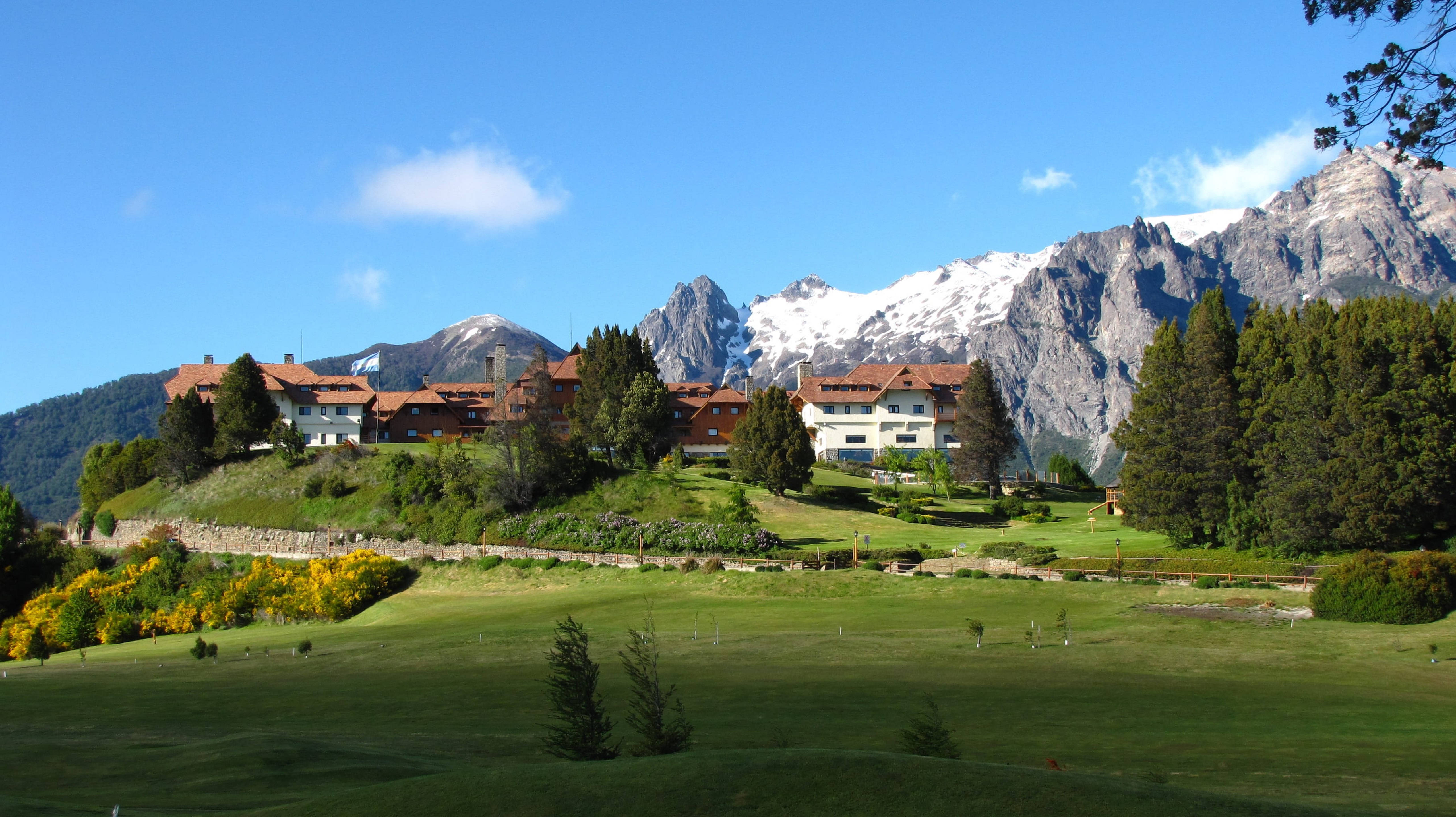 Argentina 桌布 壁紙 — 一個風景如畫的山村，紅頂房屋坐落在綠色山坡上，下方是白雪覆蓋的山峰，上方是清澈的藍天。
