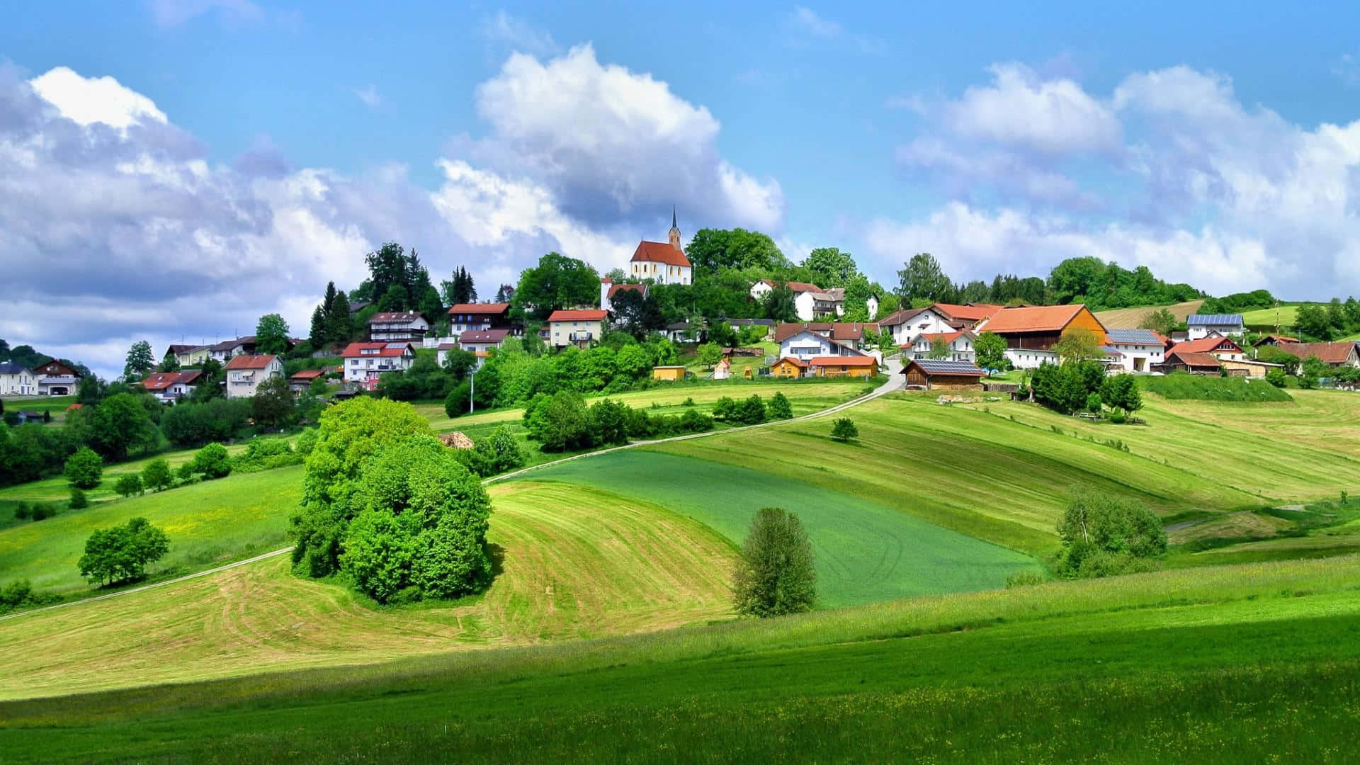 Village 桌布 壁紙 — 一個風景如畫的村莊坐落在連綿起伏的綠色山丘上，上方是明亮的藍天和蓬鬆的白雲。