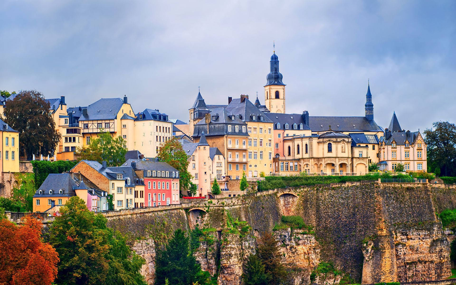 Luxembourg 桌布 壁紙 — 一個風景如畫的歐洲小鎮坐落在懸崖邊，擁有色彩繽紛的建築和一座歷史悠久的教堂，天空多雲。