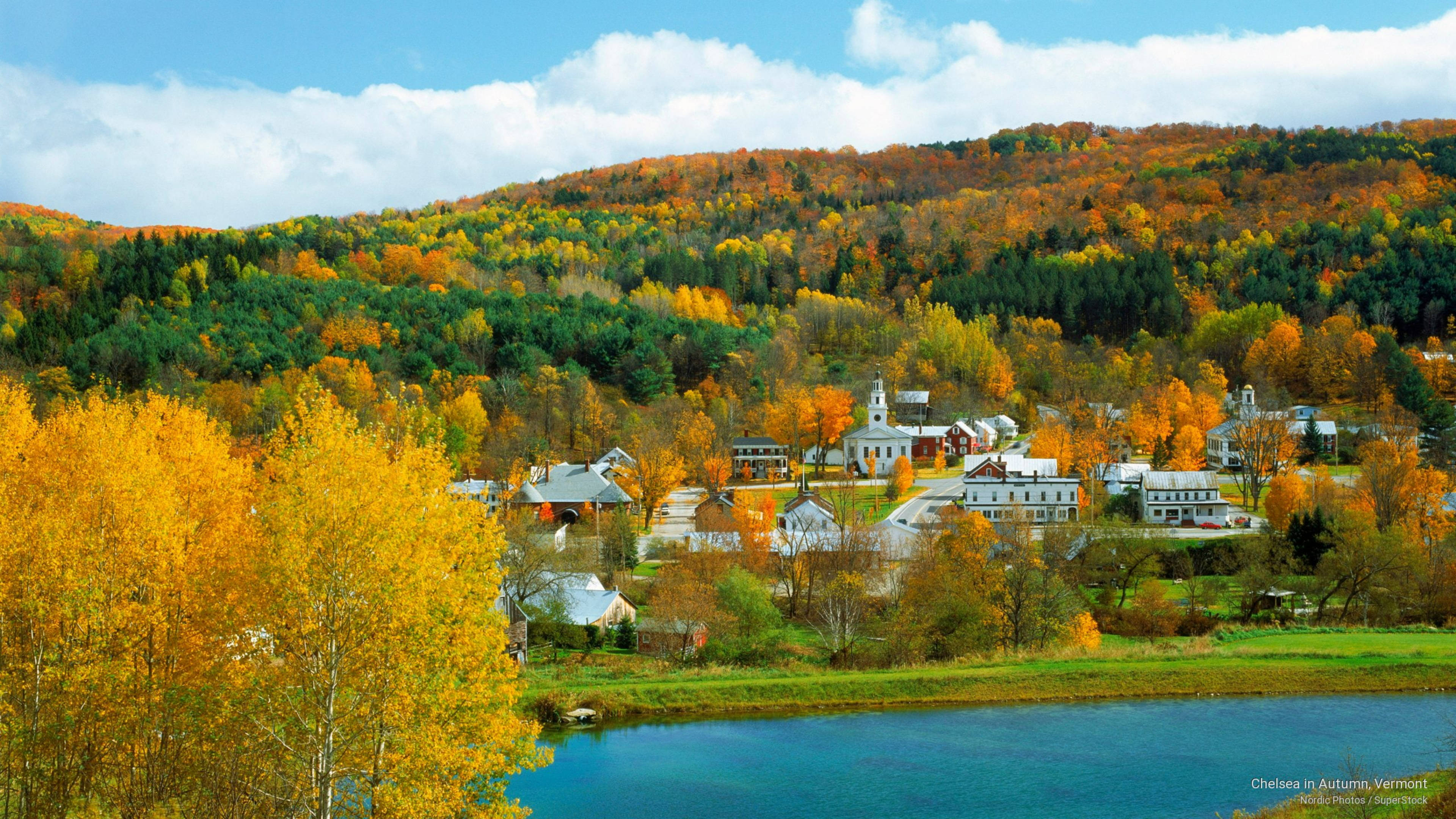 Vermont 桌布 壁紙 — 一個風景如畫的秋日村莊，坐落在充滿活力的橙色和黃色森林中，旁邊是平靜的藍色湖泊，上方是明亮的藍天和蓬鬆的白雲。