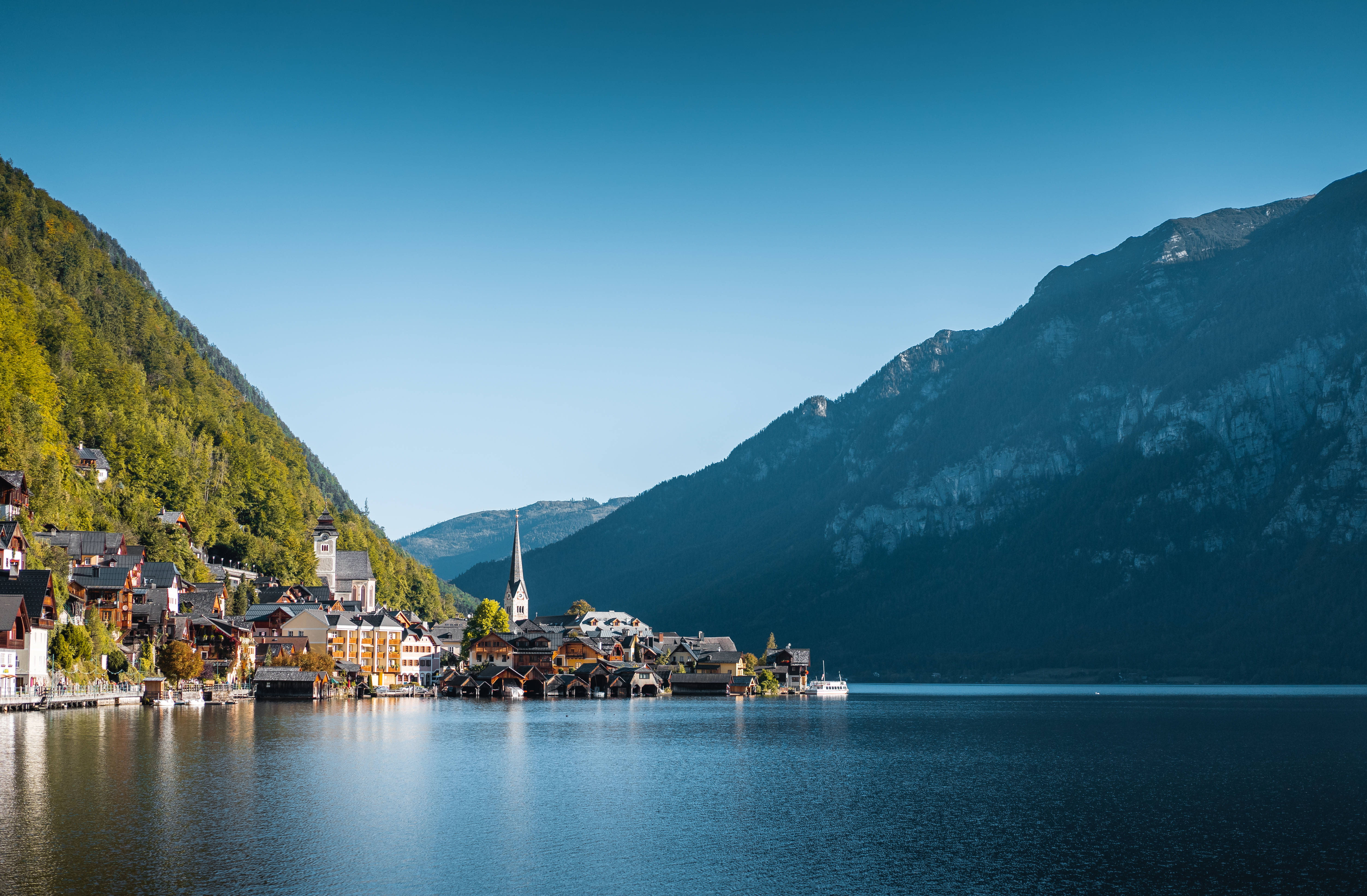 Europe 桌布 壁紙 — 一個風景如畫的阿爾卑斯山村莊依偎在寧靜的湖泊旁，四周是雄偉的綠色山脈，頭頂是清澈的藍天。