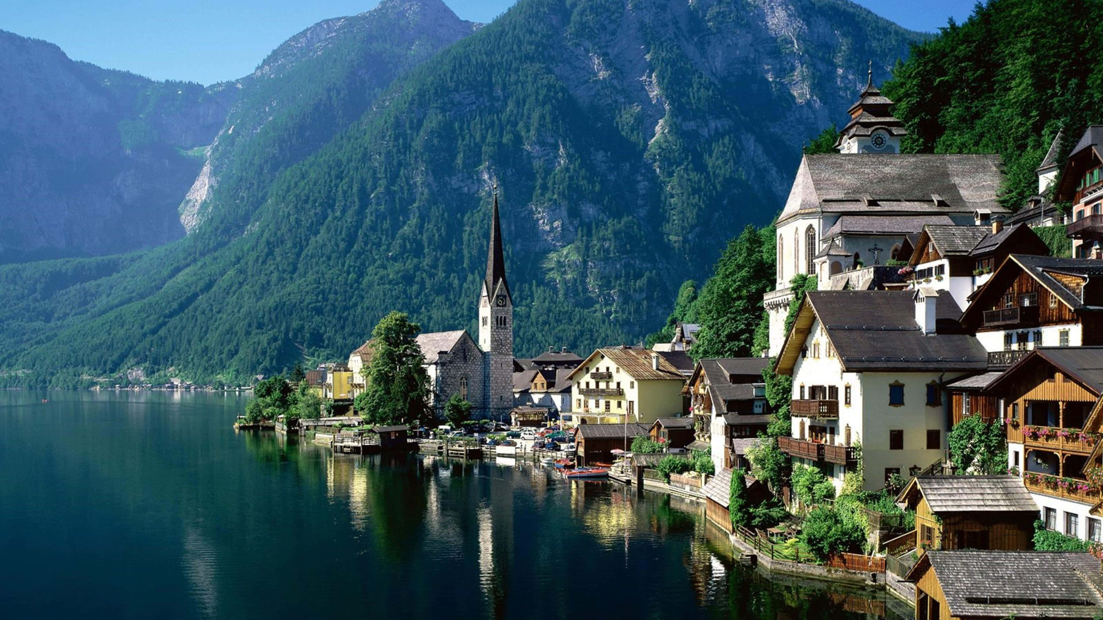 Village 桌布 壁紙 — 一個風景如畫的阿爾卑斯山村莊緊貼著寧靜的湖泊，高聳的綠色山脈在清澈的藍天下倒映在平靜的水面上。