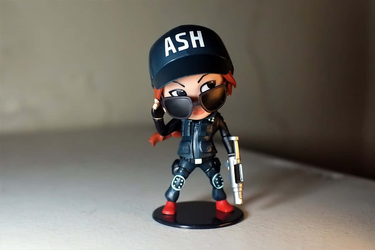 Rainbow Six Siege Ash 桌布 壁紙 — 一個風格化的Ash人偶，戴著黑色鴨舌帽、太陽眼鏡和戰鬥裝備，自信地站在展示底座上，手持武器。