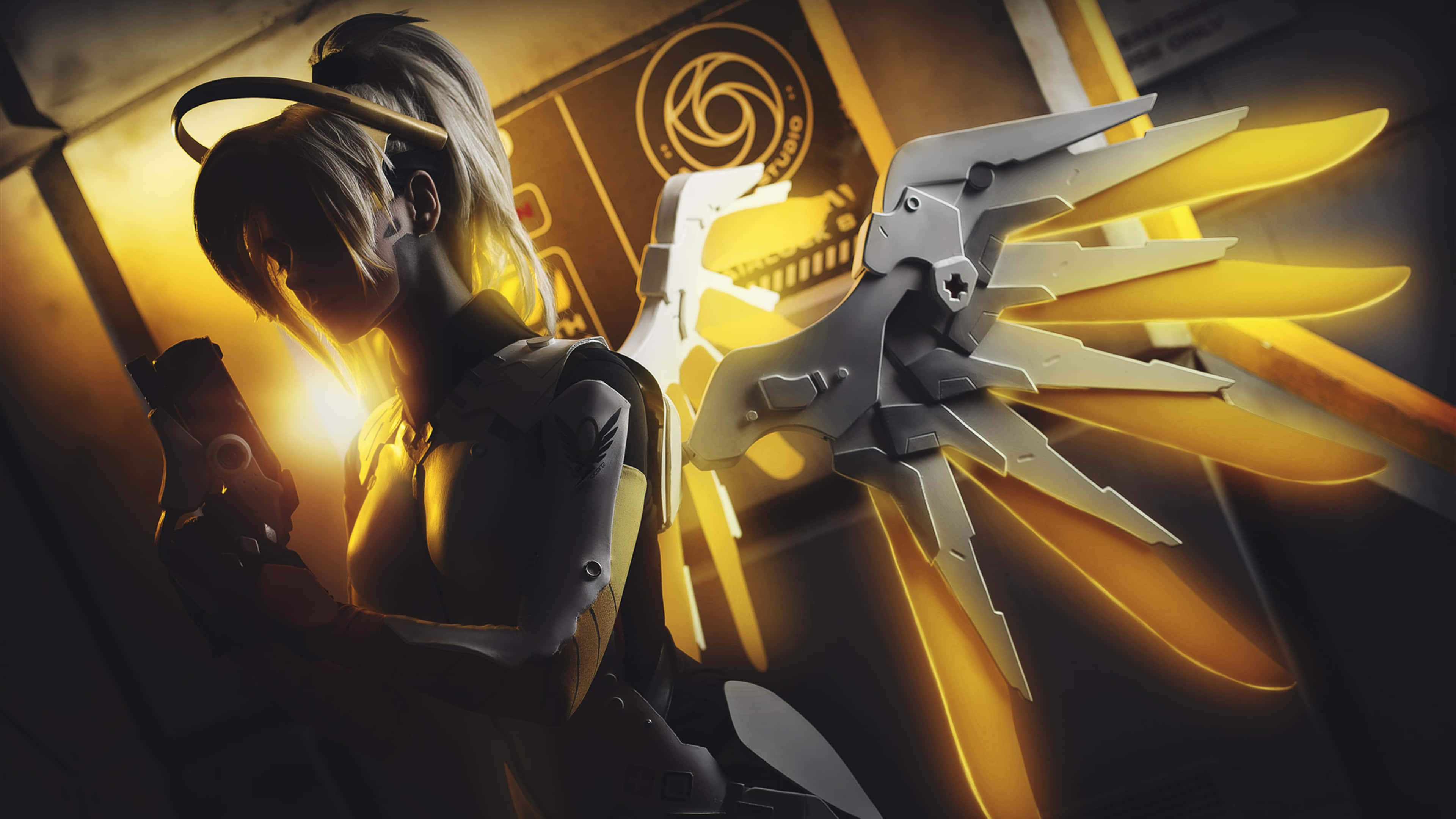 Overwatch Mercy 桌布 壁紙 — 一個風格化的動漫角色，擁有發光的金色翅膀和機械手臂，在燈光昏暗的未來感房間中手持手槍。