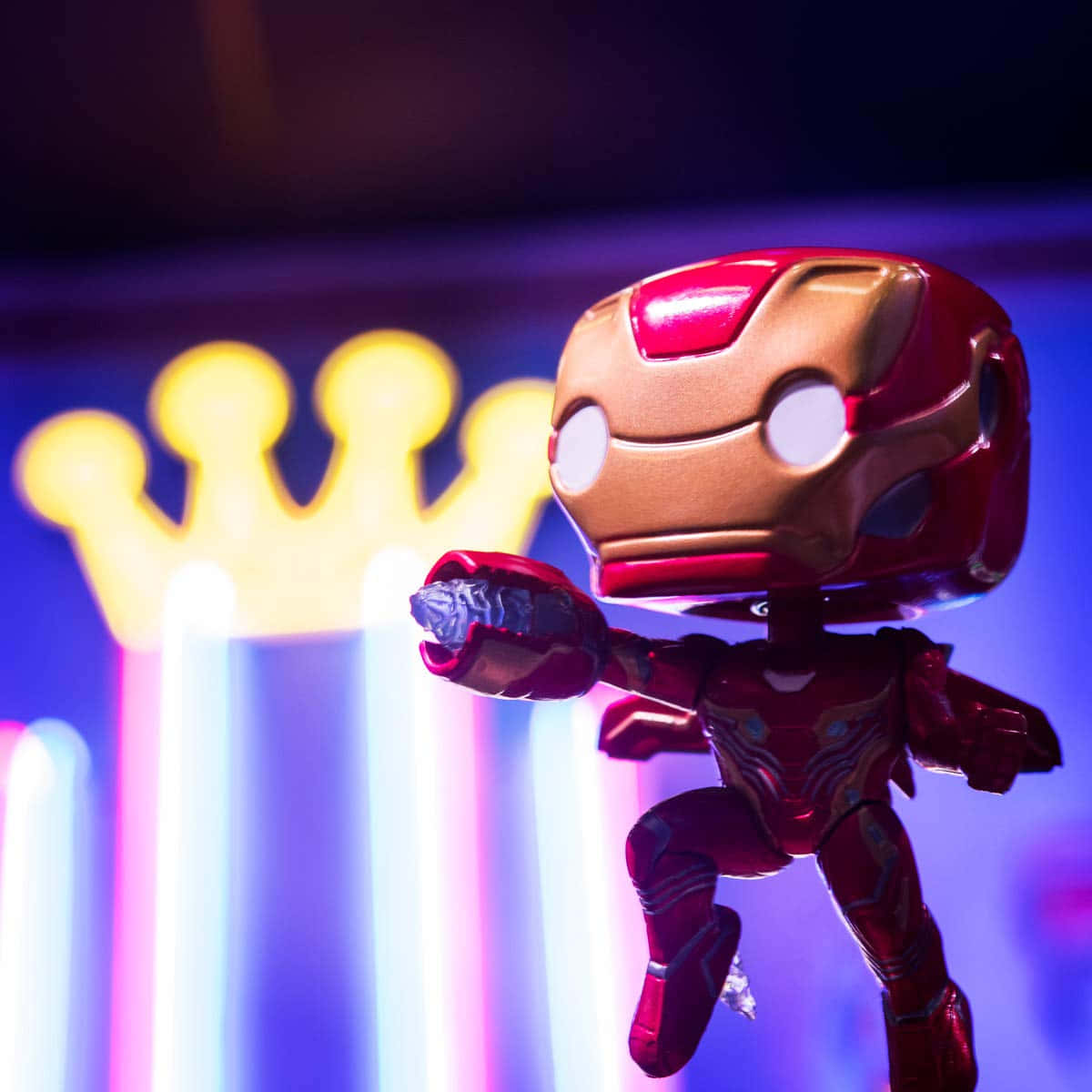 Iron Man Pop Figures 桌布 壁紙 — 一個風格化的鋼鐵人 Funko Pop 人偶在發光的霓虹皇冠背景前動態擺姿勢，帶有鮮豔的紫色和黃色燈光。