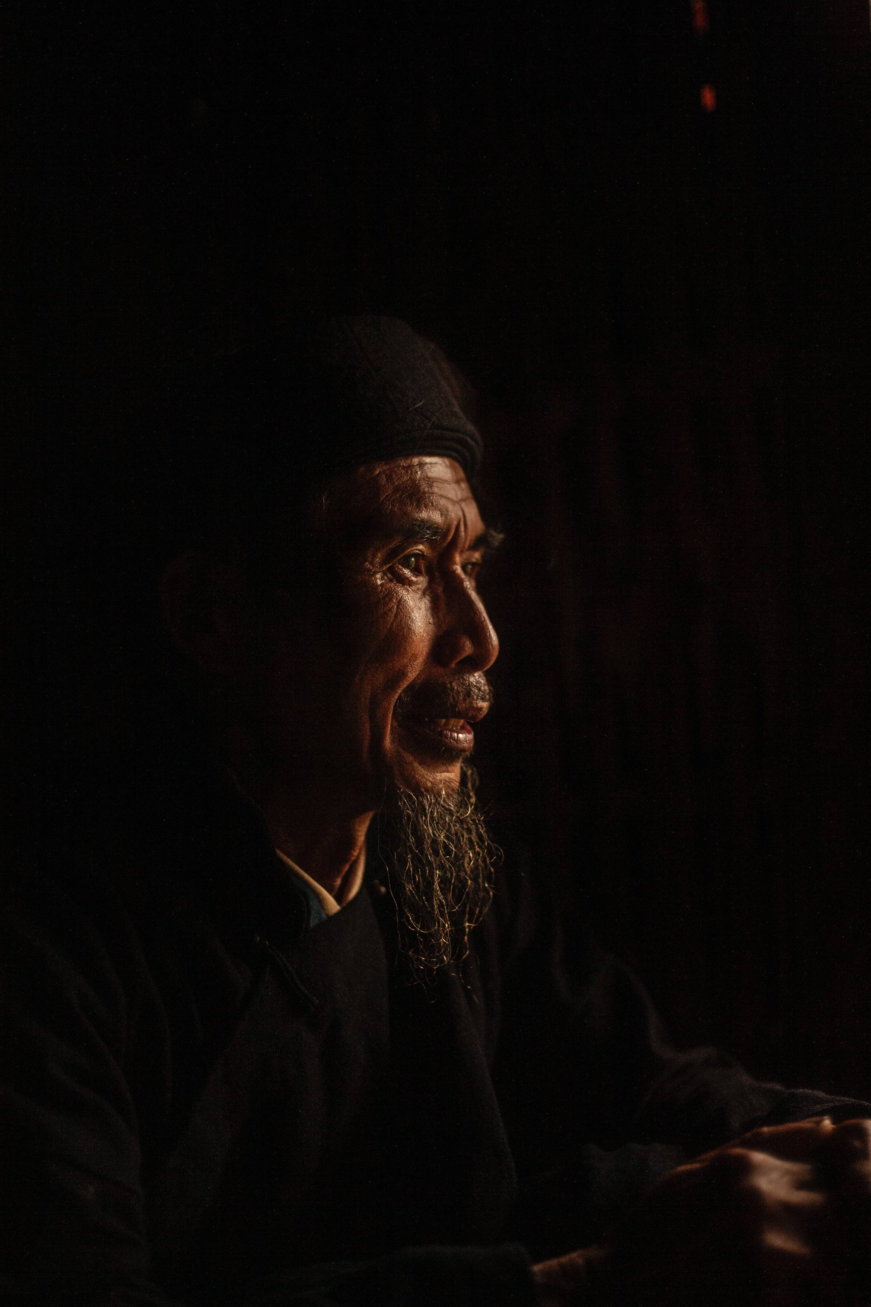 人像動態 桌布 壁紙 — 一個飽經風霜、留著長鬍鬚的男人在昏暗的光線下若有所思地凝視，他的臉被一道溫暖的聚光燈照亮，背景是黑暗的。