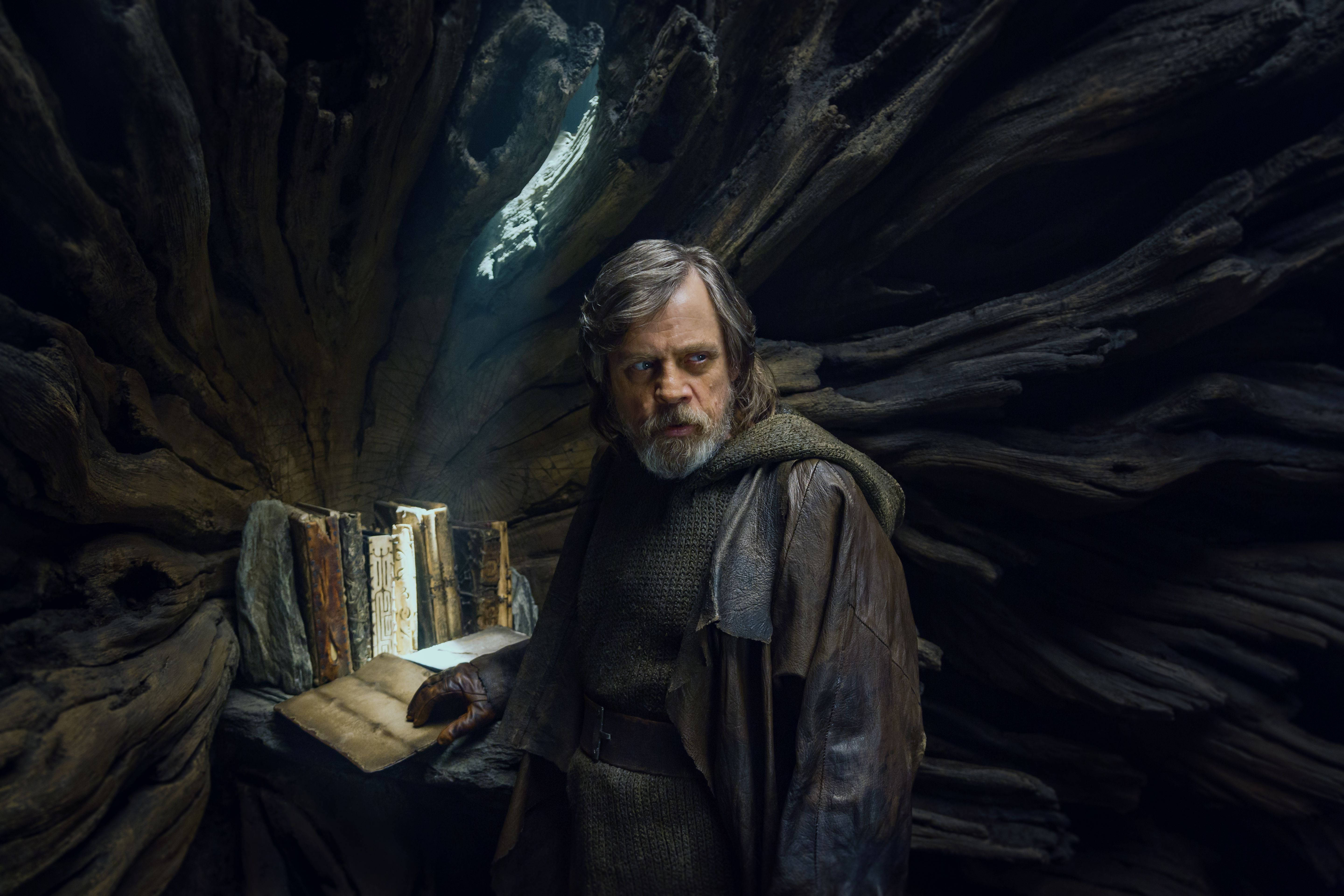 Star Wars Luke Skywalker 4K 桌布 壁紙 — 一個飽經風霜、留鬍鬚的男人站在黑暗的樹洞中，手持一本打開的書，光線從上方傾瀉而下。