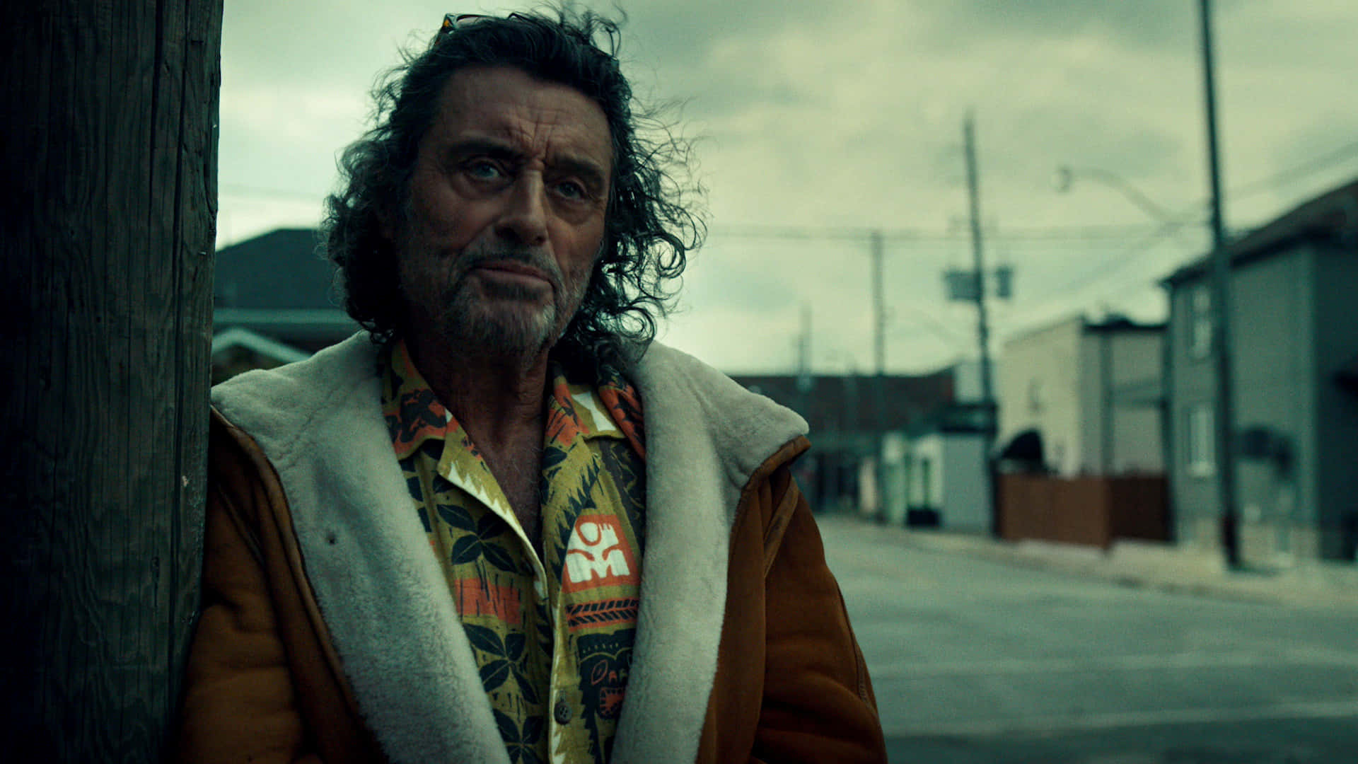 American Gods 桌布 壁紙 — 一個飽經風霜、頭髮蓬亂的男人，在灰濛濛的天空下，靠在一條荒涼街道的木柱上。
