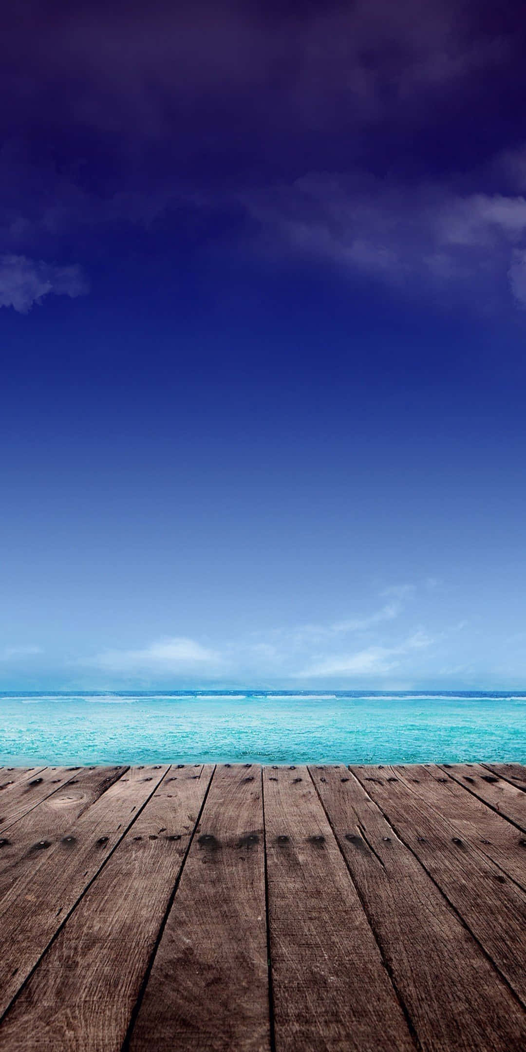 Pixel 3 Beach 桌布 壁紙 — 一個飽經風霜的木甲板俯瞰著充滿活力的綠松石色海洋，上方是深藍色的天空，點綴著幾朵雲。