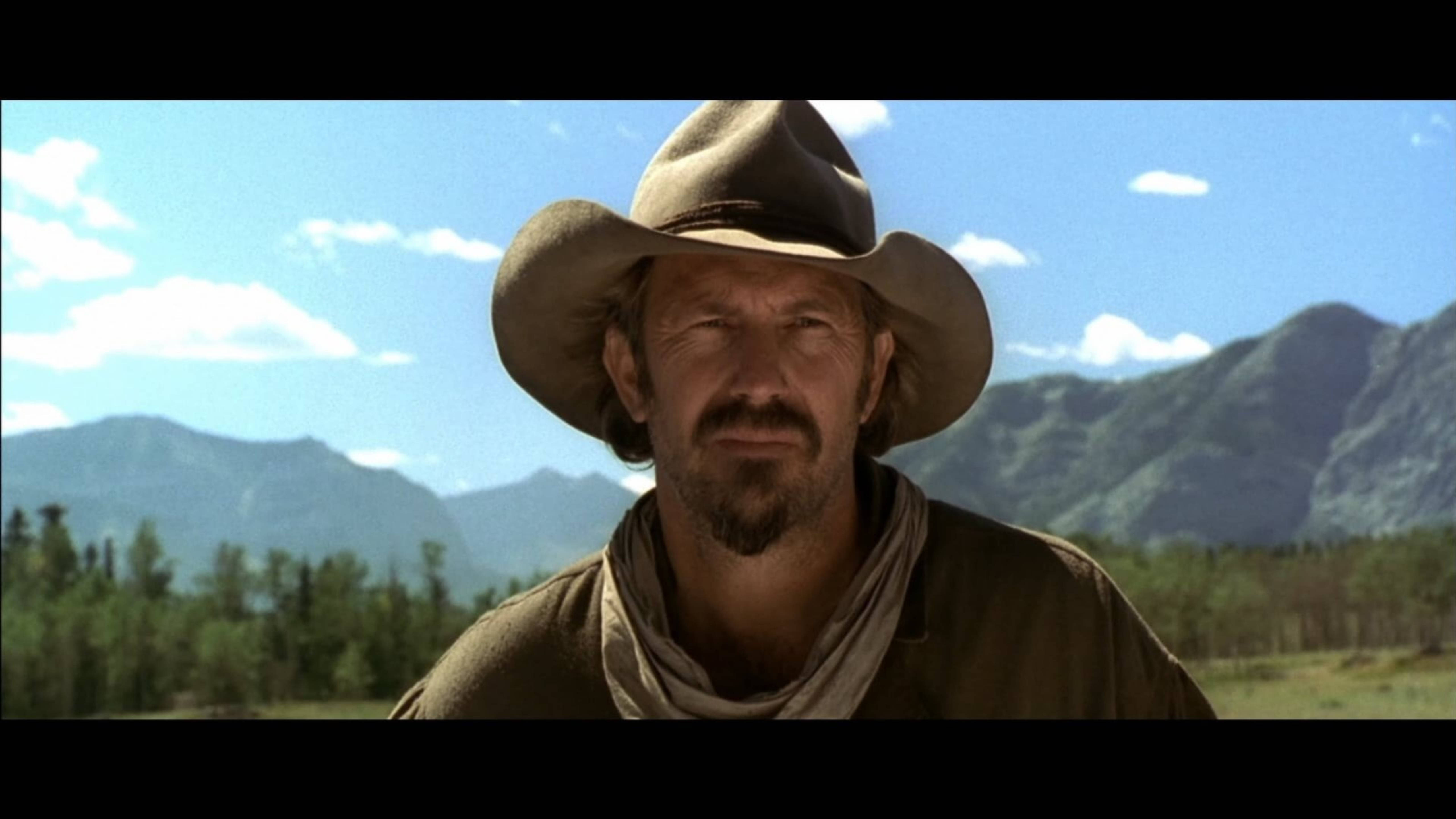 Kevin Costner 桌布 壁紙 — 一個飽經風霜的牛仔戴著寬邊帽，在遠山和明亮藍天的背景下凝視著。