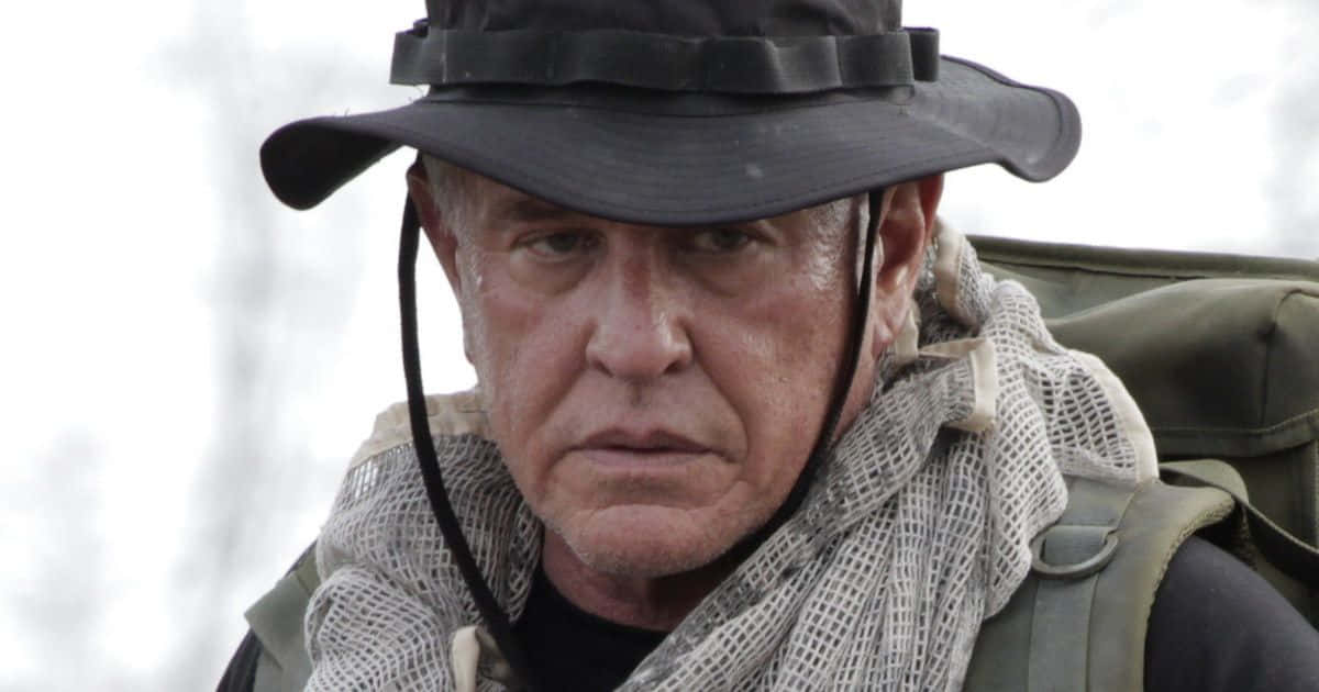 Tom Berenger 桌布 壁紙 — 一個飽經風霜的男人，戴著黑色帽子和網狀圍巾，在雪景背景下以嚴肅的表情專注地凝視。