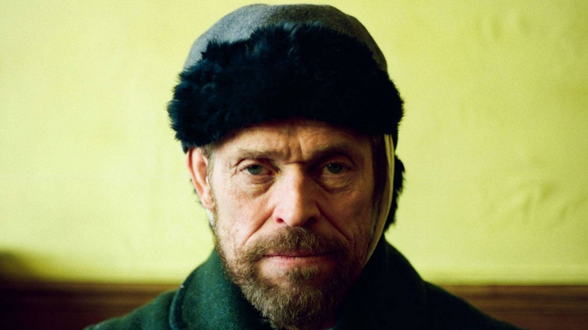 Willem Dafoe 桌布 壁紙 — 一個飽經風霜的男人，留著鬍子，戴著毛邊帽子，對著黃色牆壁凝視。
