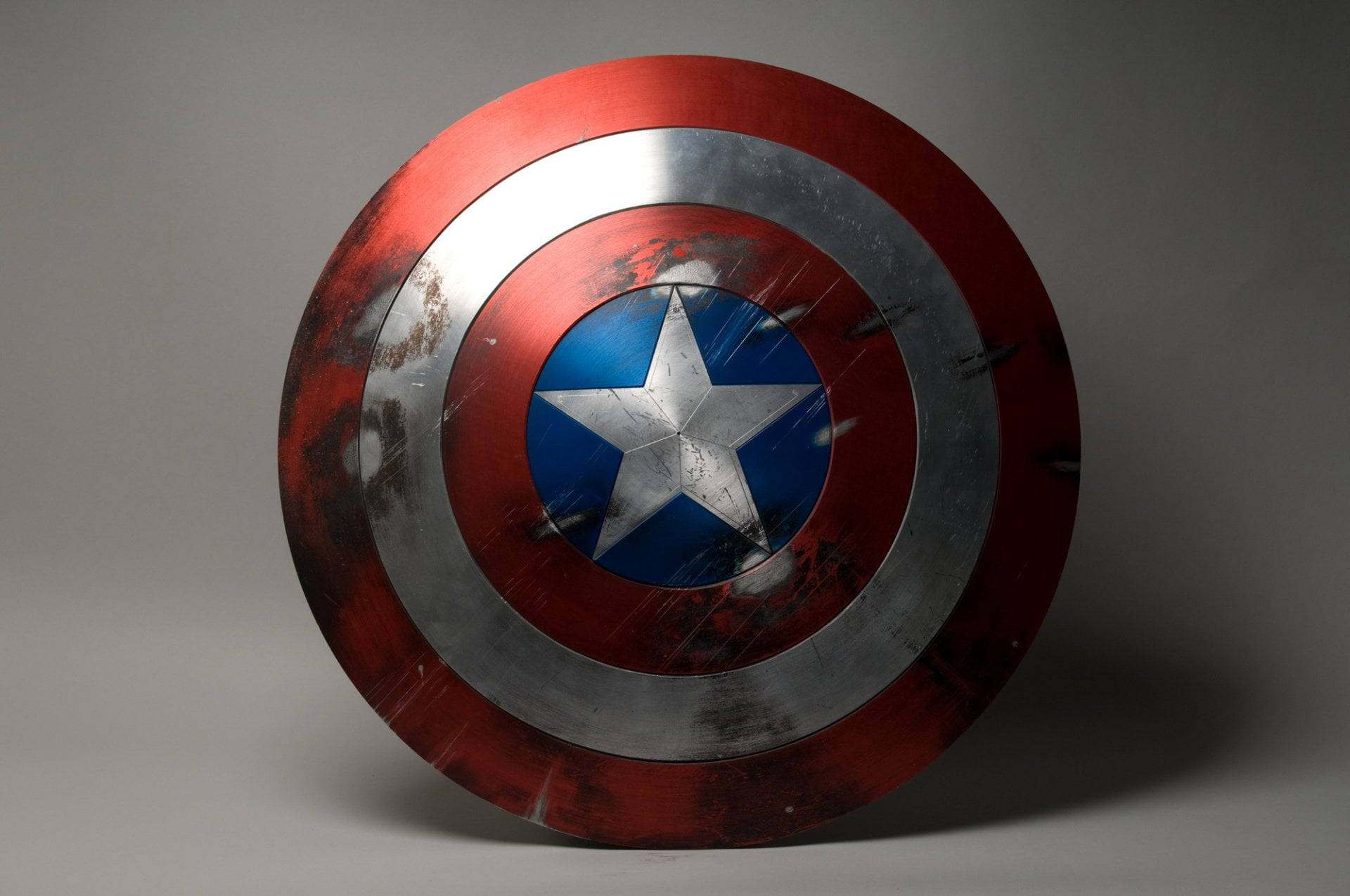 Marvel Captain America 桌布 壁紙 — 一個飽經風霜的美國隊長盾牌，帶有紅色、銀色和藍色的同心圓以及中央的星星，立在中性灰色背景前。