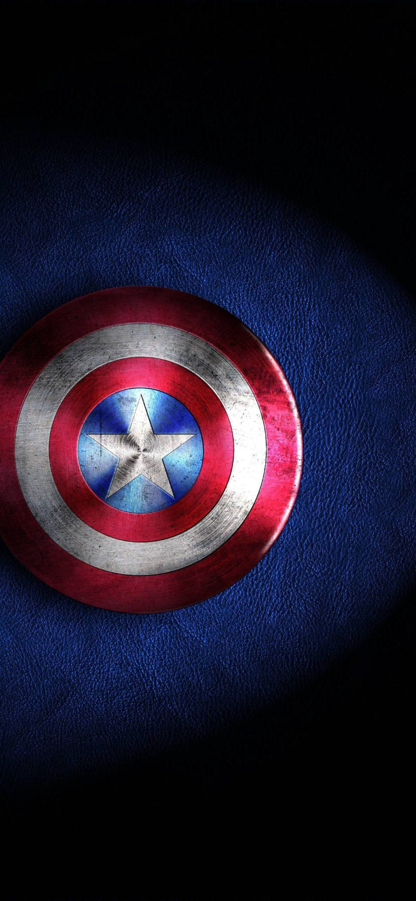 marvel iPhone xr 桌布 壁紙 — 一個飽經風霜的美國隊長盾牌放在有紋理的藍色皮革上，在黑色背景前戲劇性的聚光燈下。