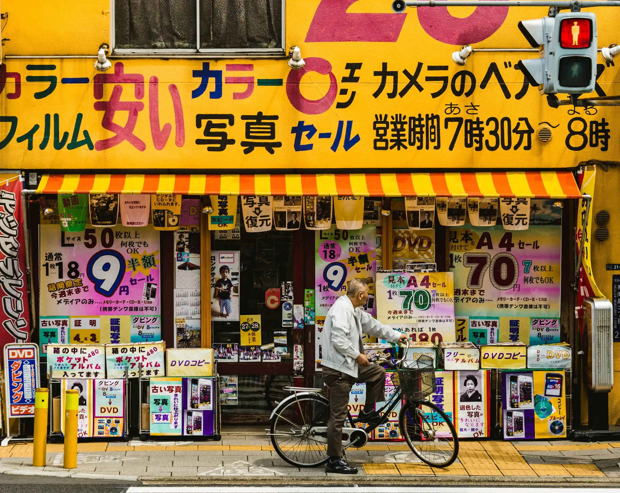 日本 桌布 壁紙 — 一個騎腳踏車的男人在充滿色彩繽紛的促銷標誌和海報的黃色日本相機店前停下來。