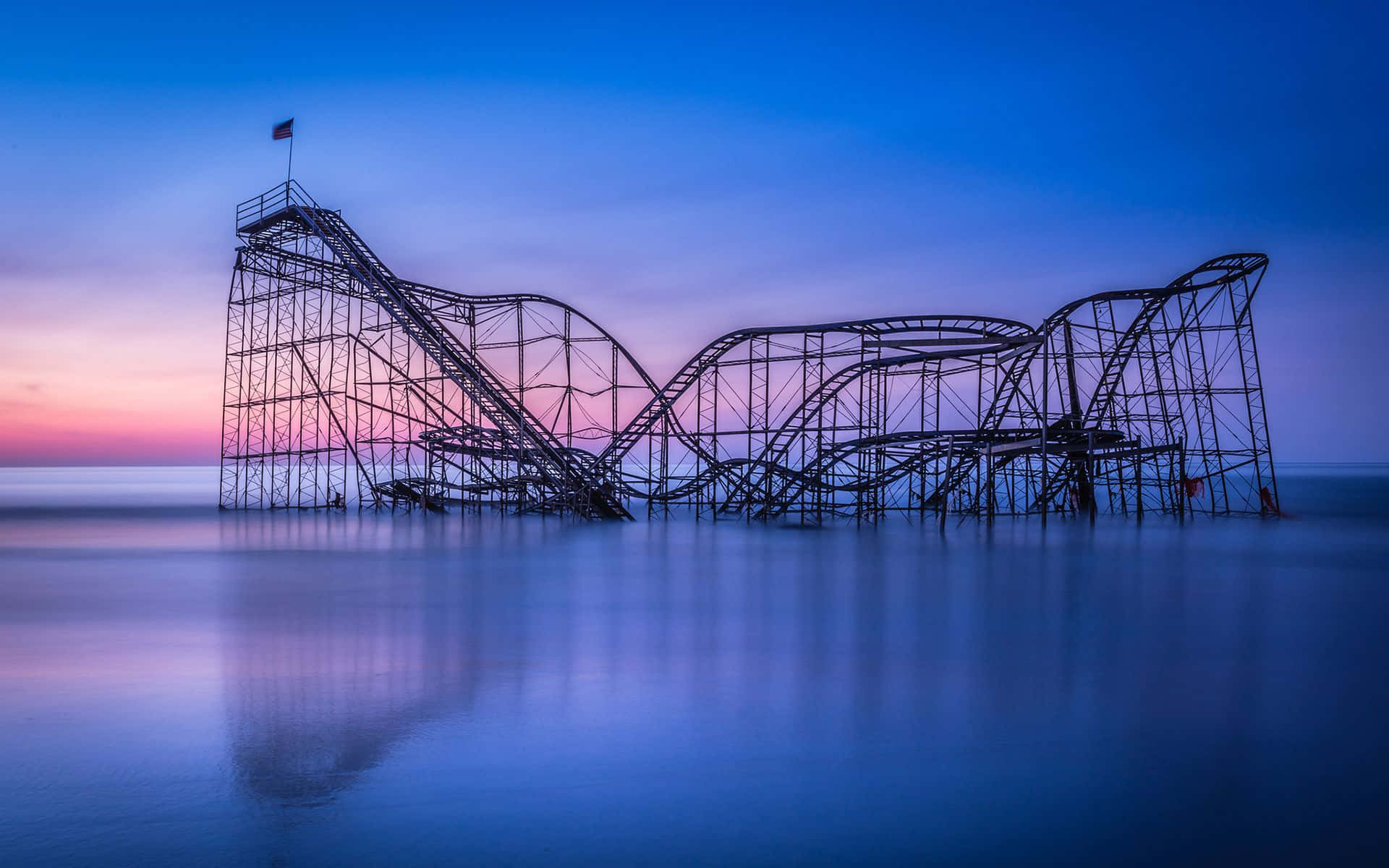 Roller Coaster 桌布 壁紙 — 一個骨架般的雲霄飛車在帶有粉紅和藍色漸層的黃昏天空下形成剪影，其倒影映照在平靜的水面上。
