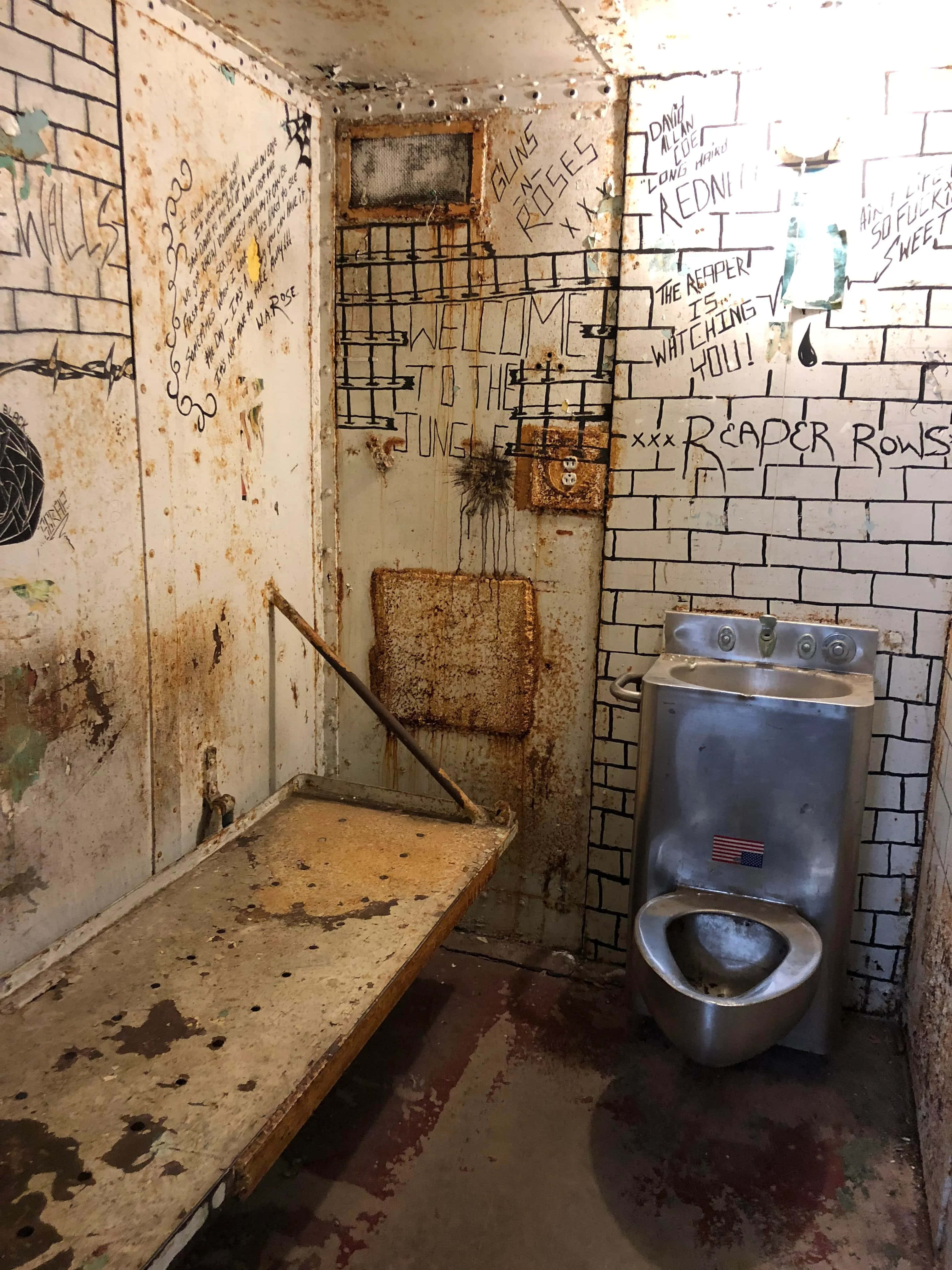 Jail Cell 桌布 壁紙 — 一個骯髒的被遺棄監獄牢房，有生鏽的金屬裝置、塗鴉覆蓋的牆壁，以及在閃爍燈光下的一張生鏽長凳。