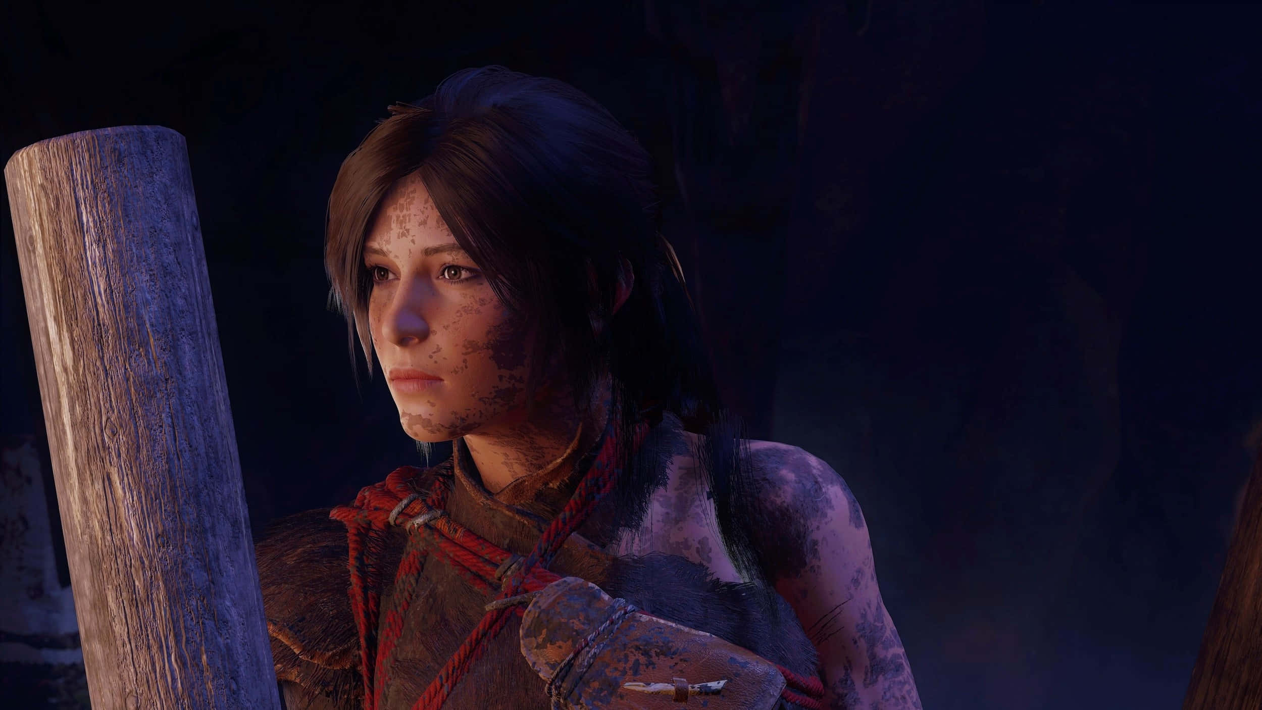 1440p Shadow Of The Tomb Raider 桌布 壁紙 — 一個骯髒的電玩角色，臉上沾滿污垢，身穿紅色繩索盔甲，站在昏暗陰影環境中的木柱旁。