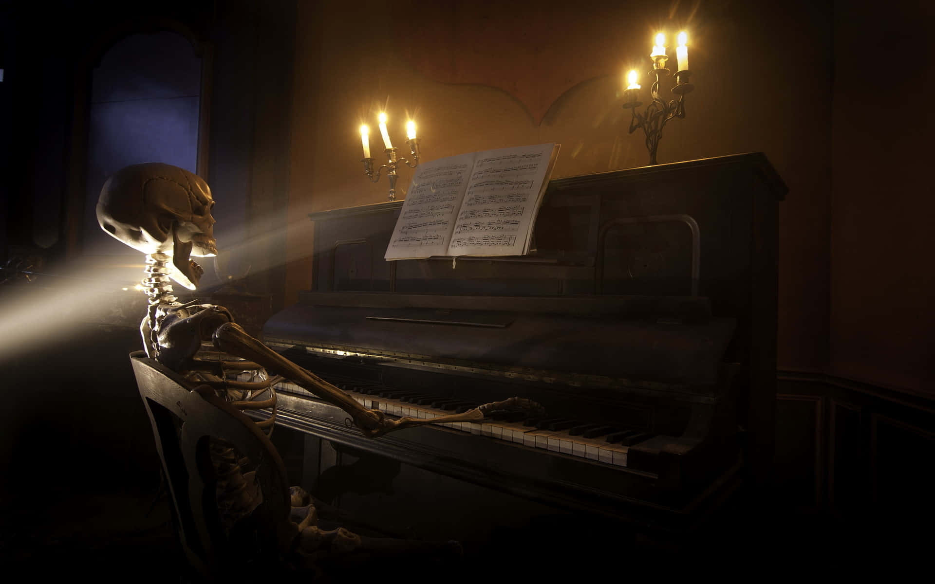 Halloween Music 桌布 壁紙 — 一個骷髏坐在昏暗房間的三角鋼琴前，被燭光和一束陽光照亮。