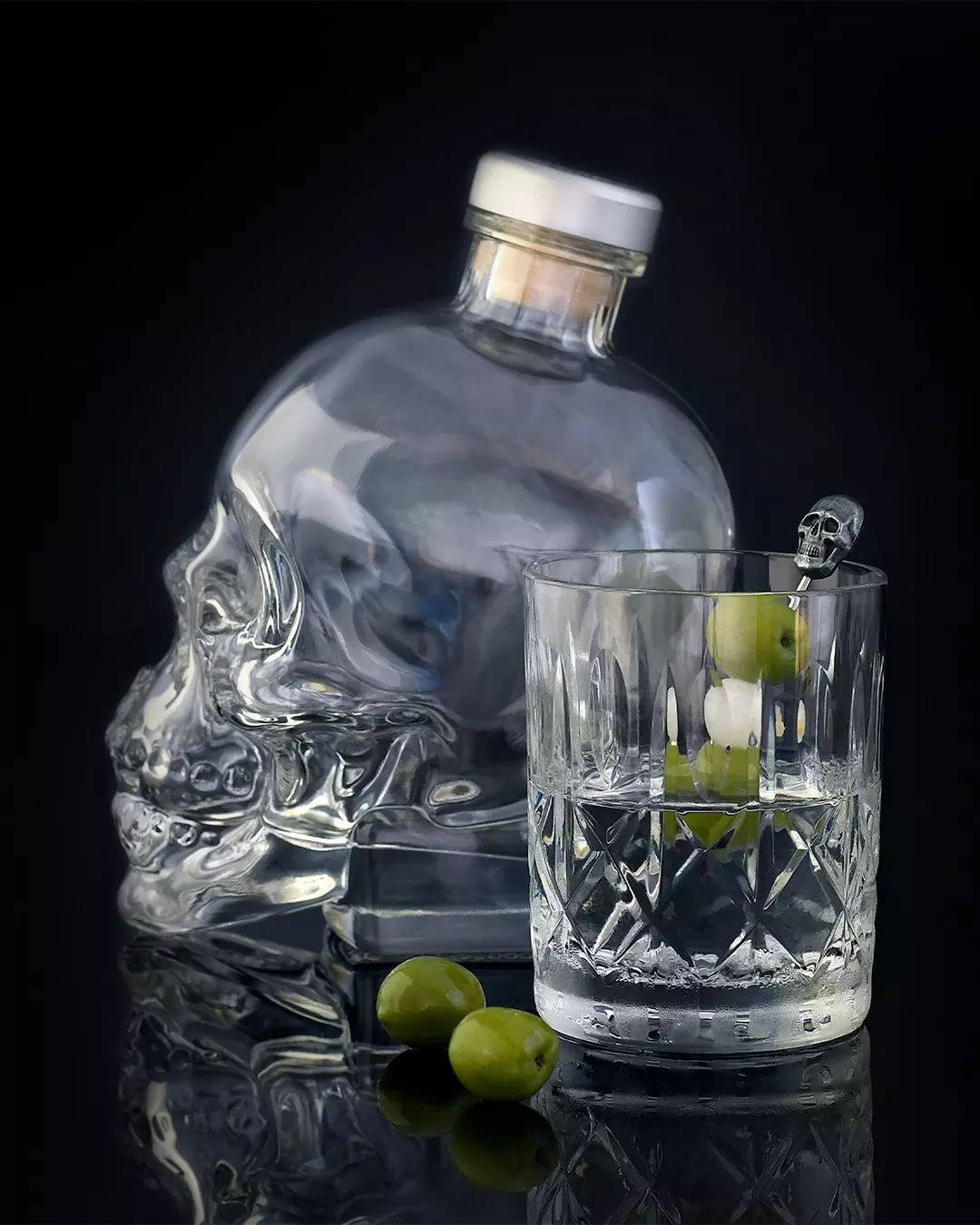 Drink 桌布 壁紙 — 一個骷髏形狀的玻璃伏特加酒瓶旁邊放著一個水晶杯，裡面有橄欖和一個骷髏攪拌棒，放在反光的黑色表面上。
