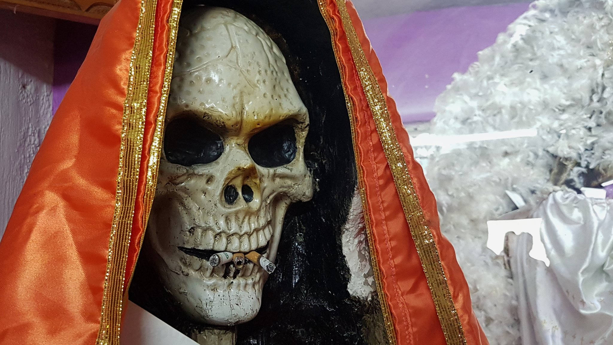 Santa Muerte 桌布 壁紙 — 一個骷髏頭穿著橙色連帽長袍，飾有金色邊飾，咬著一支香煙，背景是白色水晶。