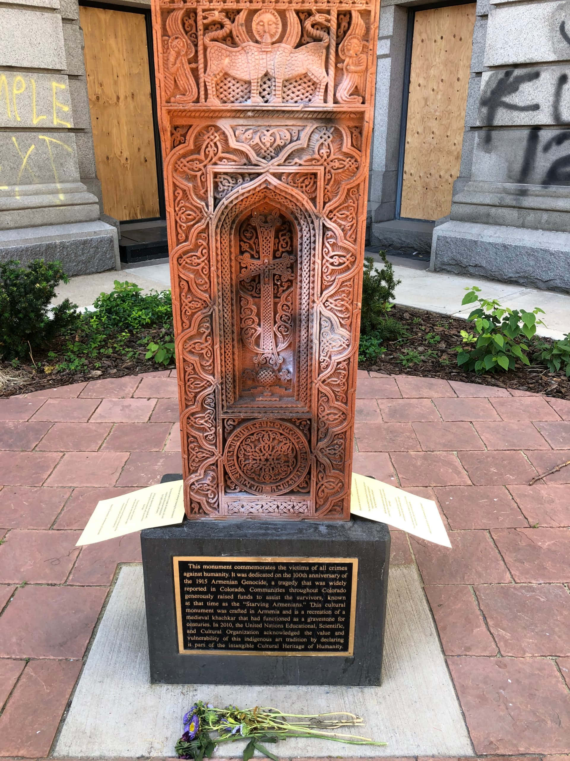 Memorial 桌布 壁紙 — 一個高大、雕刻精緻的木製亞美尼亞紀念十字架矗立在石基座上，位於鋪砌的庭院中，附近建築物上有塗鴉。