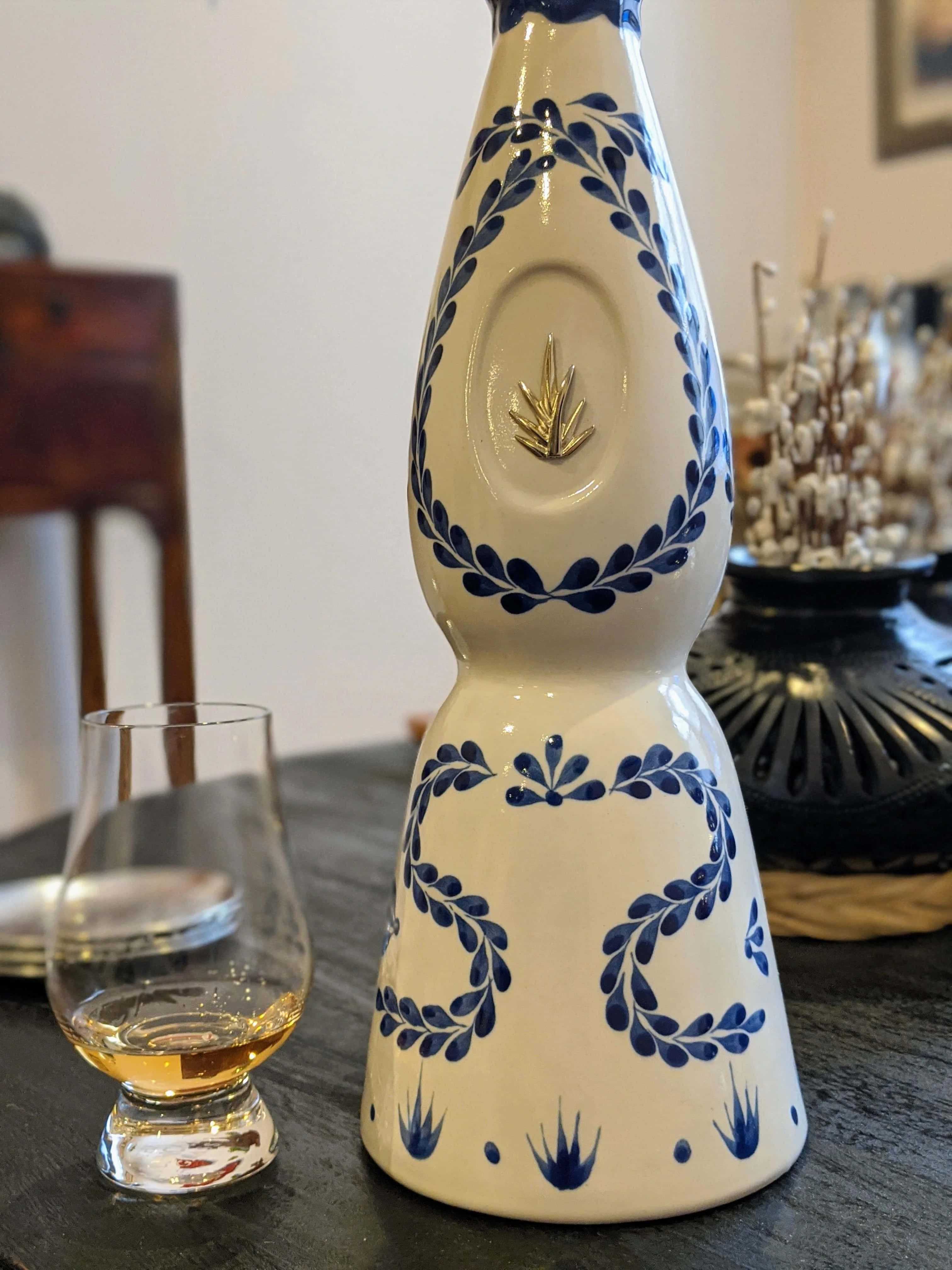 alcohol 桌布 壁紙 — 一個高大的白色陶瓷瓶，帶有藍色葉子圖案和金色葉子徽章，放在一個深色木桌上，旁邊是一杯琥珀色液體。