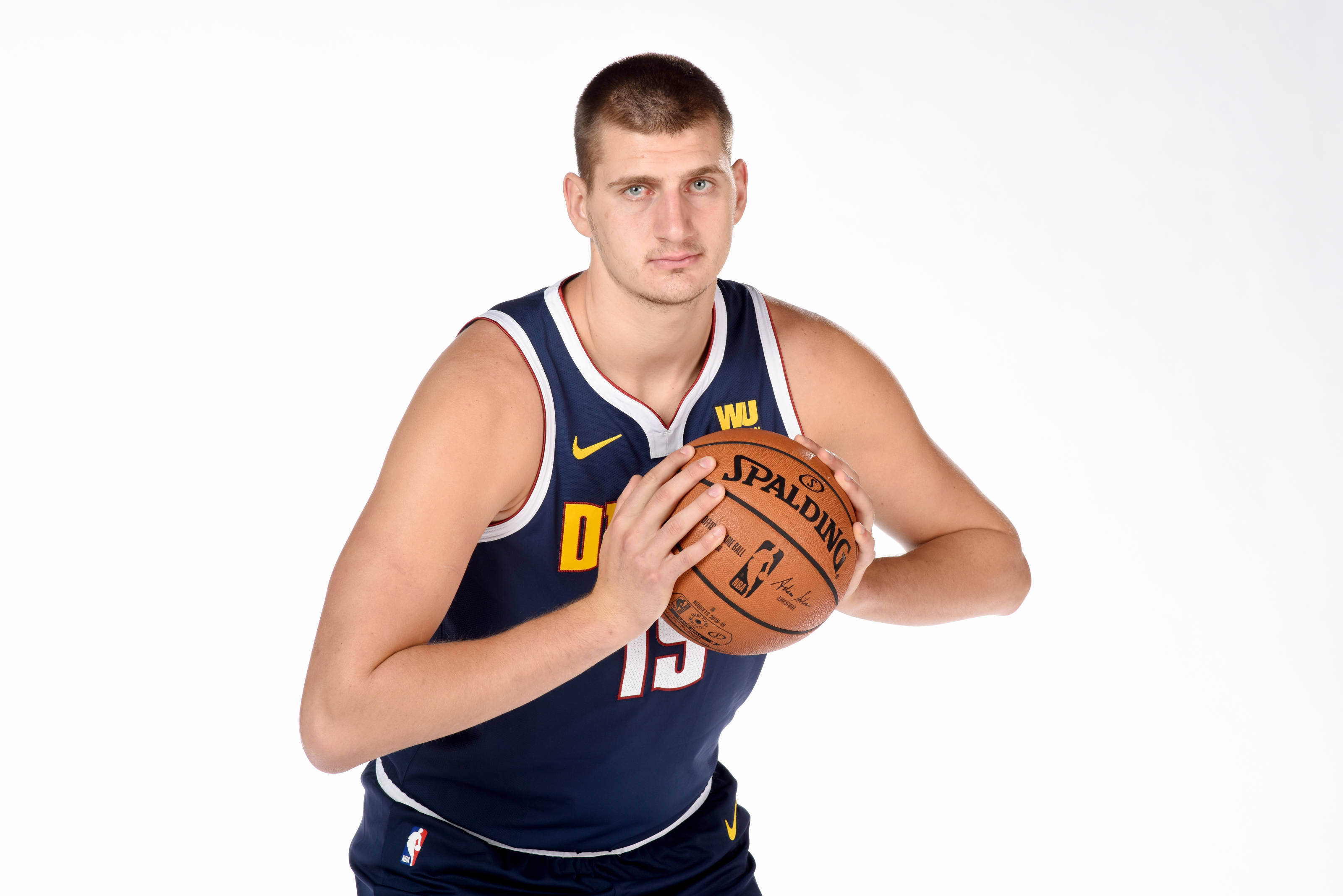 Jokic 桌布 壁紙 — 一個高大的籃球運動員穿著海軍藍球衣，在純白色背景前拿著一個Spalding籃球。
