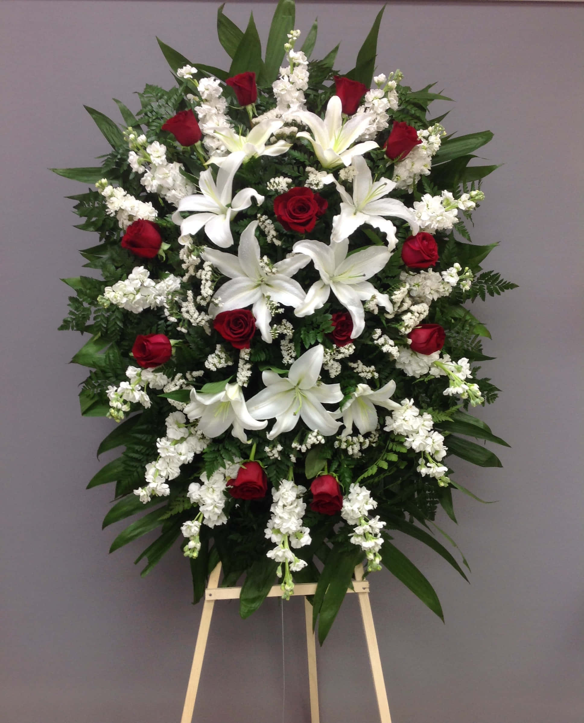 Funeral Flower Arrangements 桌布 壁紙 — 一個高大的花卉佈置，包括白色百合、紅玫瑰和綠葉，展示在木製畫架上，背景是灰色牆壁。