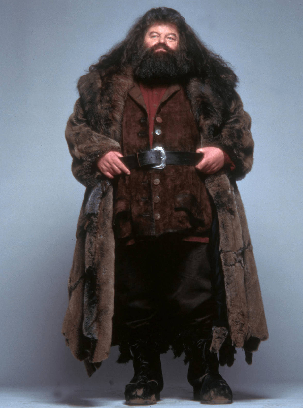 rubeus hagrid 桌布 壁紙 — 一個高大的鬍鬚男，身穿破舊的棕色皮草大衣，自信地站在純灰色工作室背景前。
