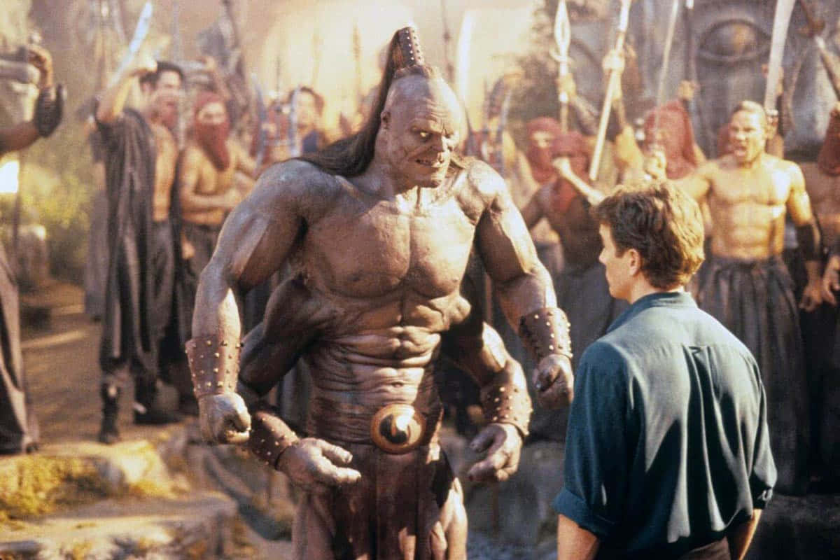 Mortal Kombat Goro 桌布 壁紙 — 一個高大肌肉發達、頭髮尖刺的外星戰士，在陽光明媚、擠滿武裝旁觀者的外星市場中與一名人類男子對峙。