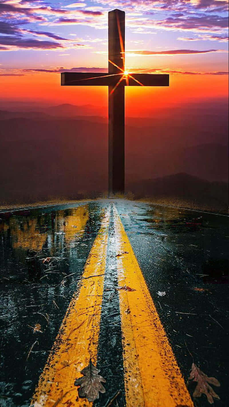 Jesus Saves 桌布 壁紙 — 一個高聳的十字架矗立在一條濕漉漉的道路盡頭，天空是充滿活力的日落，帶有橙色和紫色的雲彩。