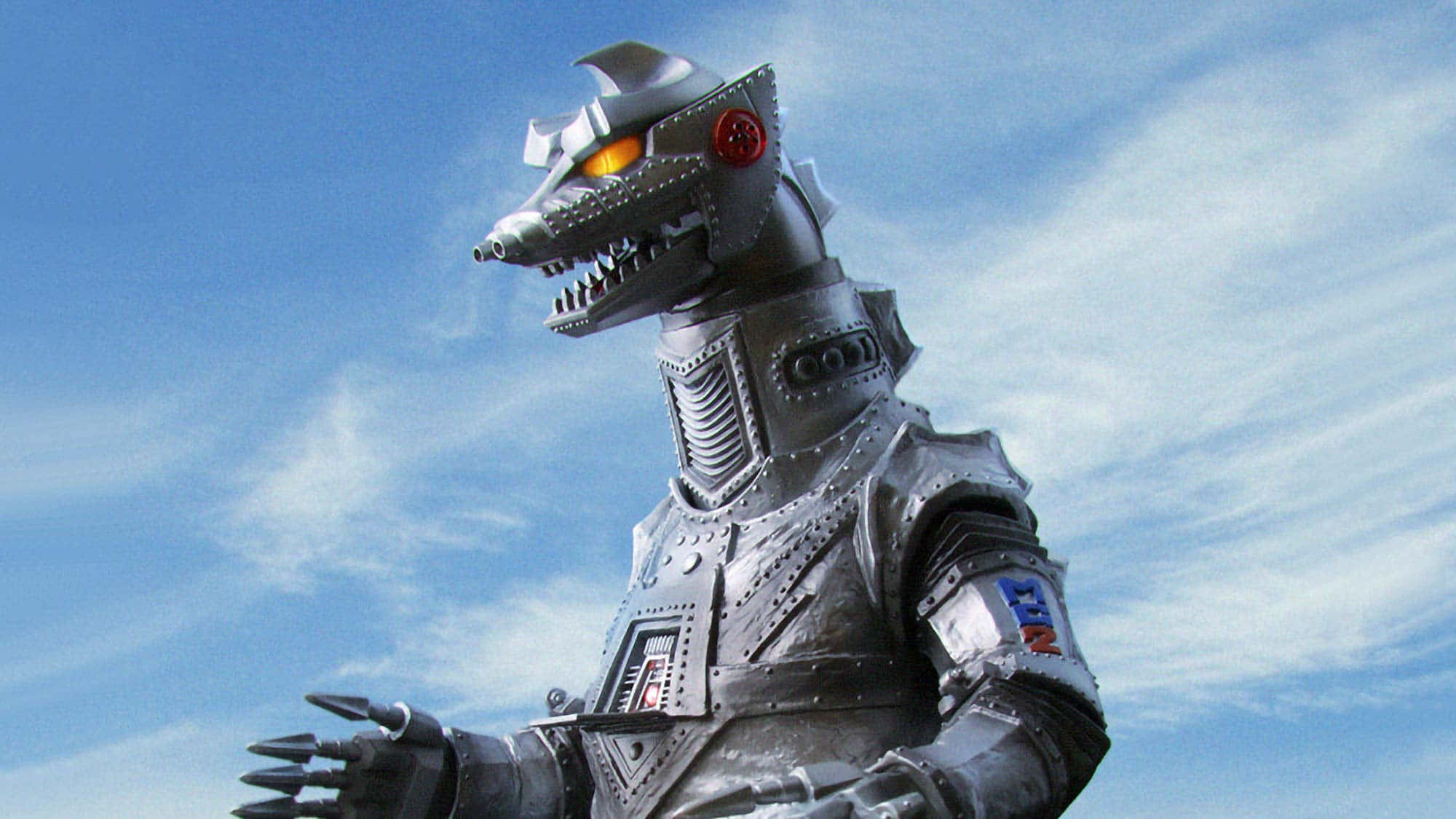 Robot Godzilla 桌布 壁紙 — 一個高聳的銀色機器人怪物，擁有發光的橙色眼睛和鋒利的牙齒，站在有縷縷白雲的明亮藍天下。